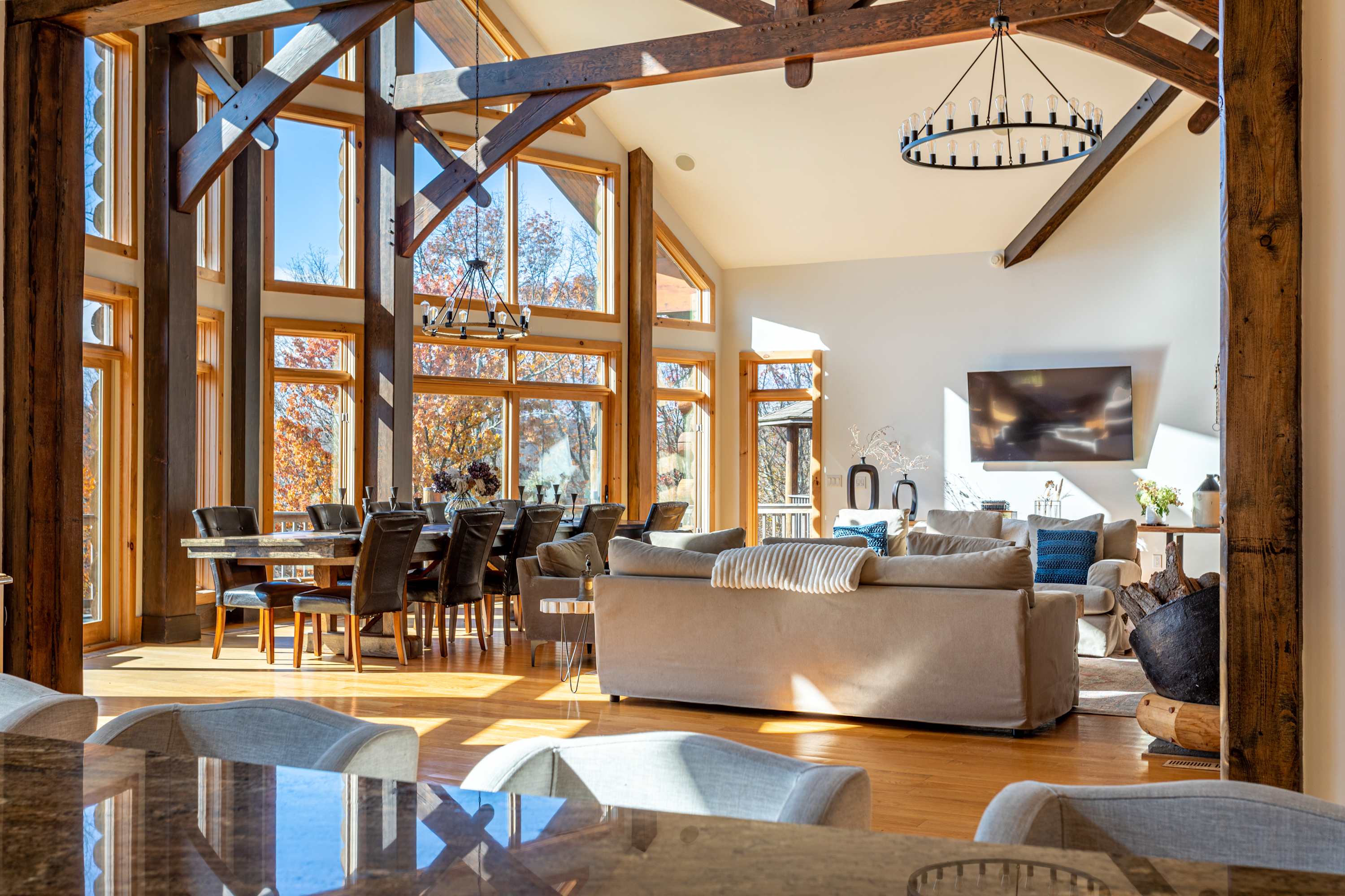 A bright, open-concept living and dining space showcases a long wooden dining table with dark chairs, surrounded by expansive floor-to-ceiling windows offering views of autumn trees. A large chandelier hangs above, and a comfortable living area with sofas is visible in the foreground.