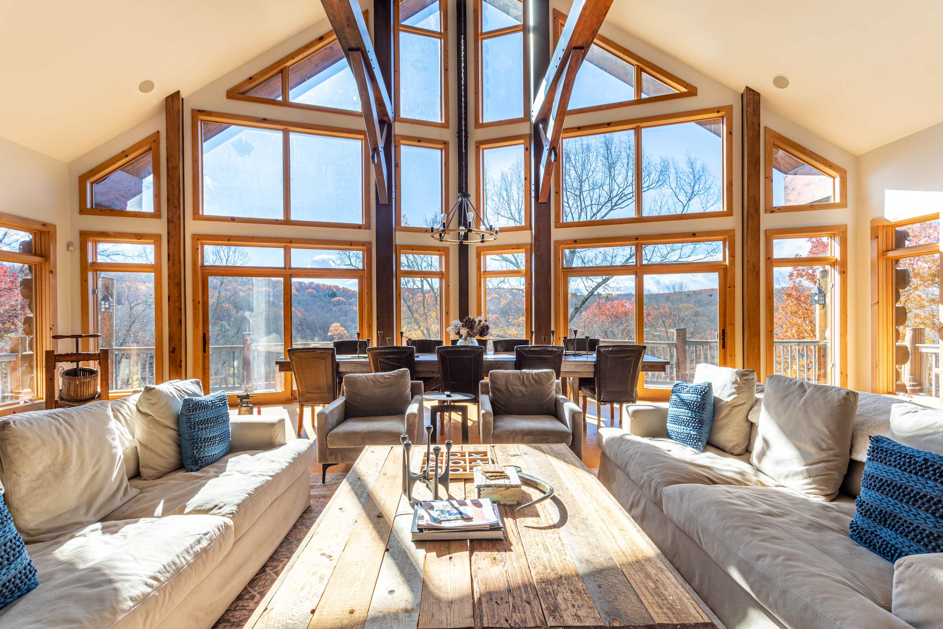 Expansive living area with soaring vaulted ceilings and floor-to-ceiling windows offering panoramic views of the surrounding forest, featuring two large light-colored sofas and a rustic wooden coffee table. A dining area is visible in the background.