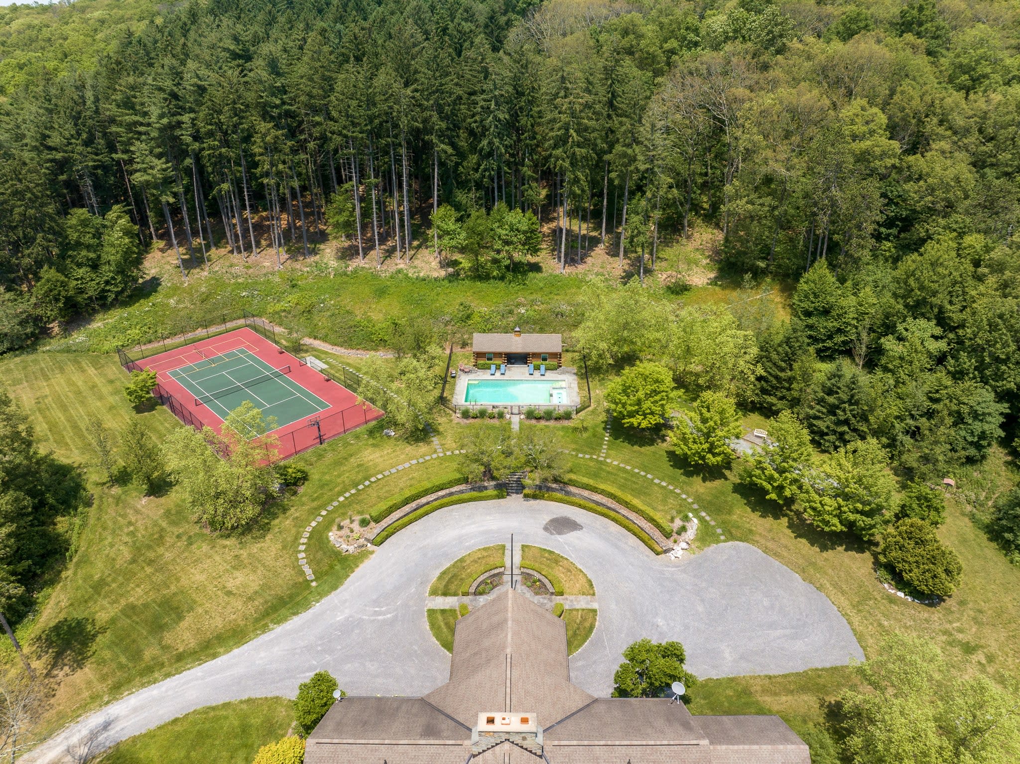 An aerial view showcasing the property's amenities, including a large circular driveway leading to the main house, a swimming pool with a log cabin pool house, and a red tennis court, all nestled within a vast forested landscape.