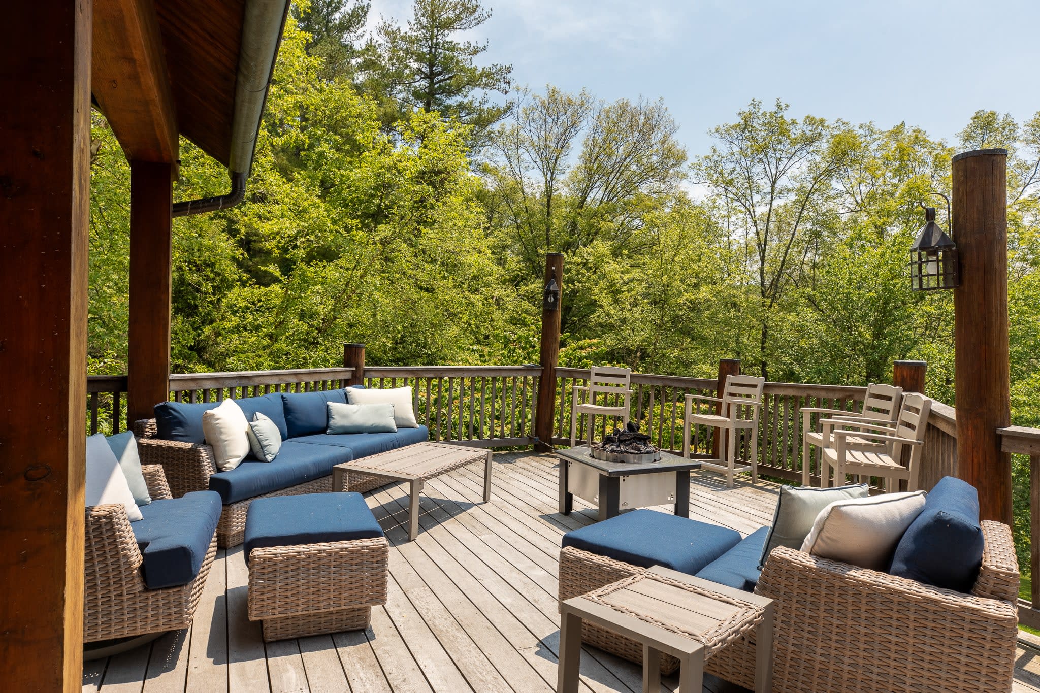 An open-air section of the wooden deck, surrounded by dense green trees, features a large blue-cushioned outdoor sectional sofa and a separate seating area with a fire pit table. The deck is made of light-colored wood planks.