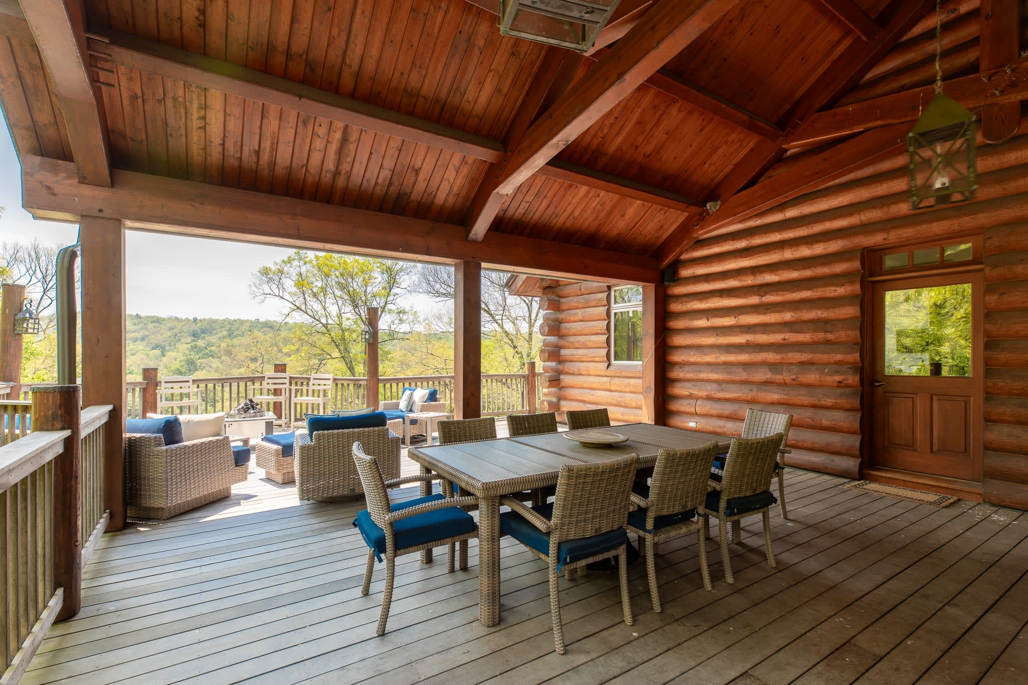 A spacious covered outdoor deck with a high wooden ceiling and exposed beams, featuring a large wicker dining table with blue-cushioned chairs and a separate lounge area with wicker sofas and armchairs. The deck overlooks a lush green valley.
