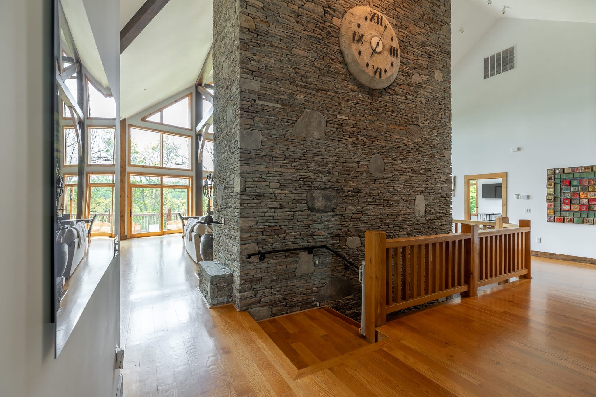 Grand interior space with a towering stone wall featuring a large decorative clock, polished hardwood floors, and a wooden railing leading to a lower level. Large windows offer views of the outdoors.