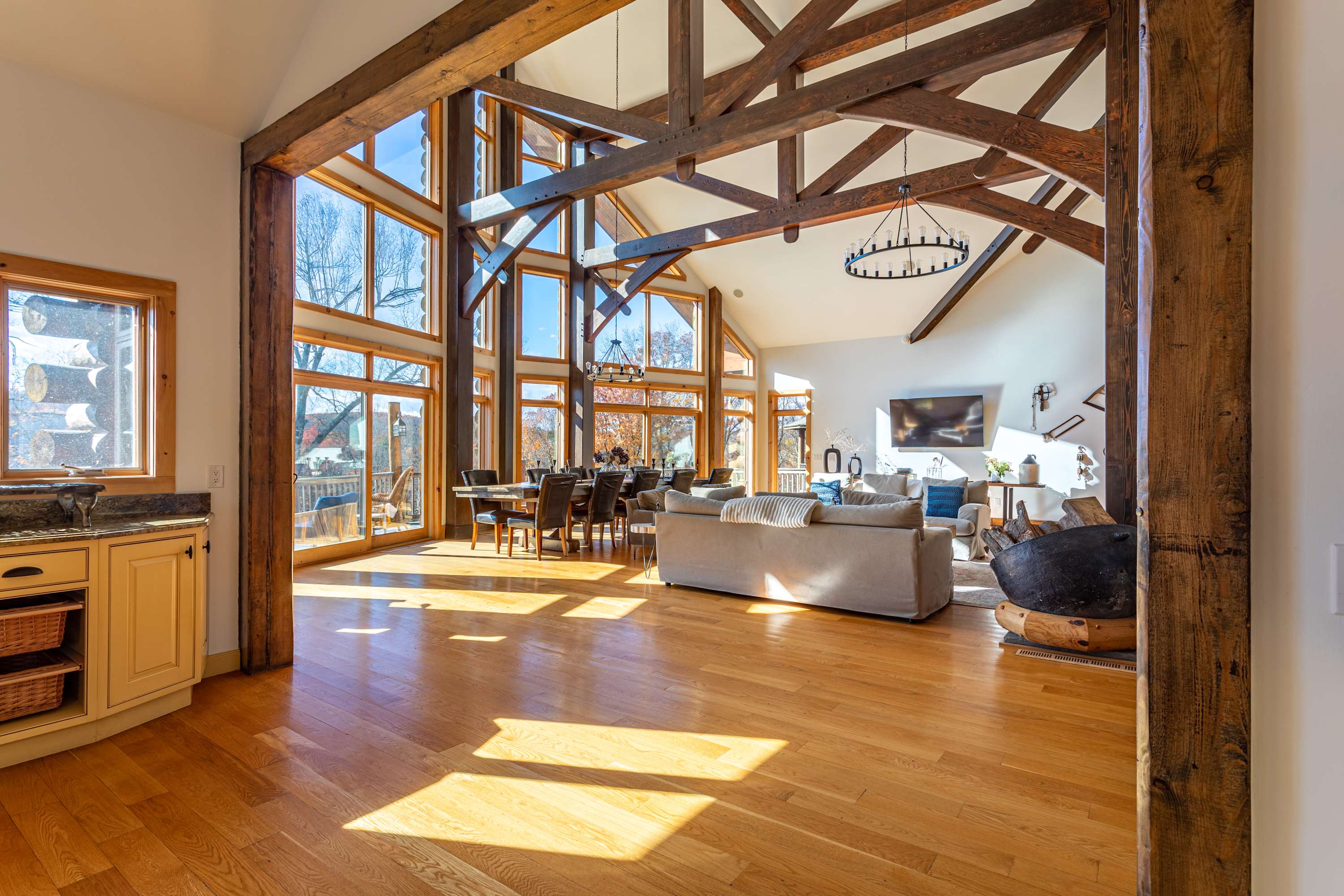 An expansive open-concept living and dining area featuring a massive stone fireplace, exposed timber beams, and floor-to-ceiling windows offering abundant natural light and outdoor views.