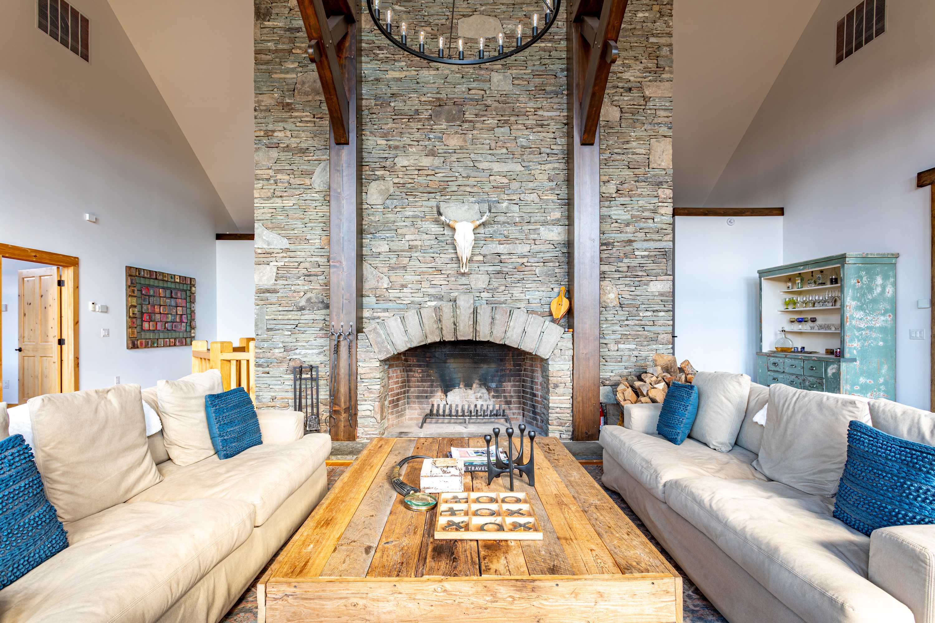 A cozy living room centered around a grand stone fireplace, featuring two comfortable sofas with blue accent pillows, a rustic wooden coffee table, and exposed wooden beams overhead.