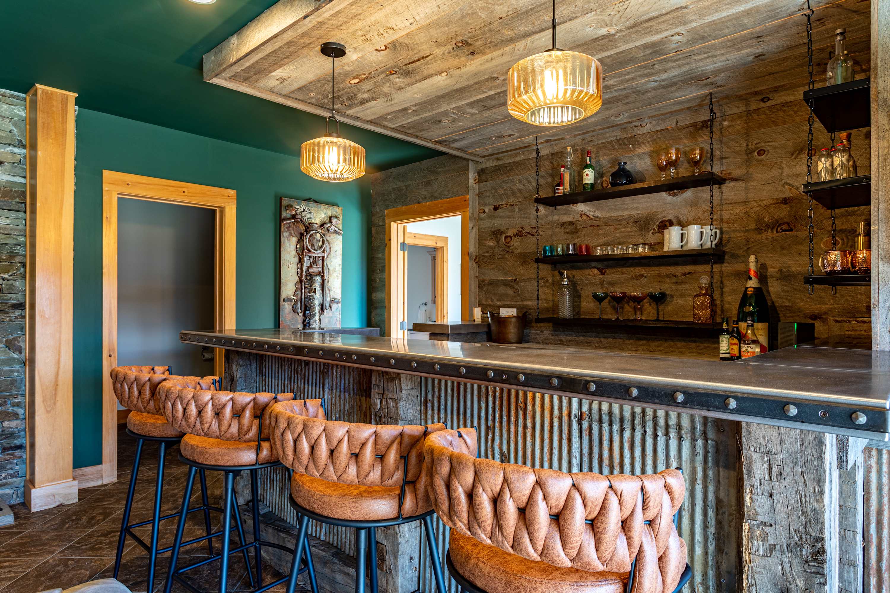 A wide shot of the rustic bar, featuring a metal countertop and five brown leather bar stools, set against a reclaimed wood wall with open shelves. The ceiling above the bar is also reclaimed wood, contrasting with the green painted ceiling of the wider room.