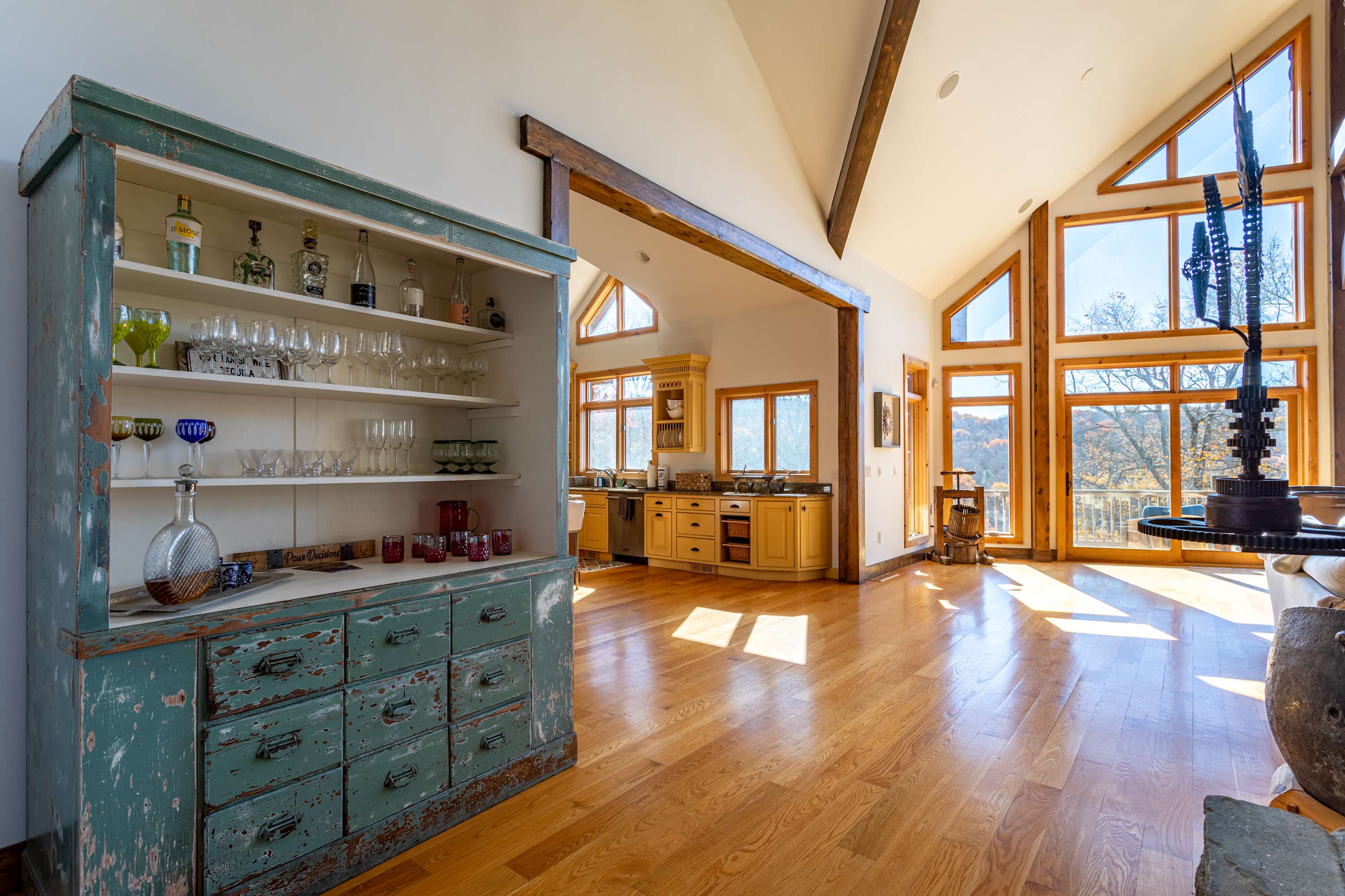 A wide view of the rustic kitchen with light yellow cabinets, granite countertops, and a distressed teal hutch, opening into a spacious living area with large windows and hardwood floors.
