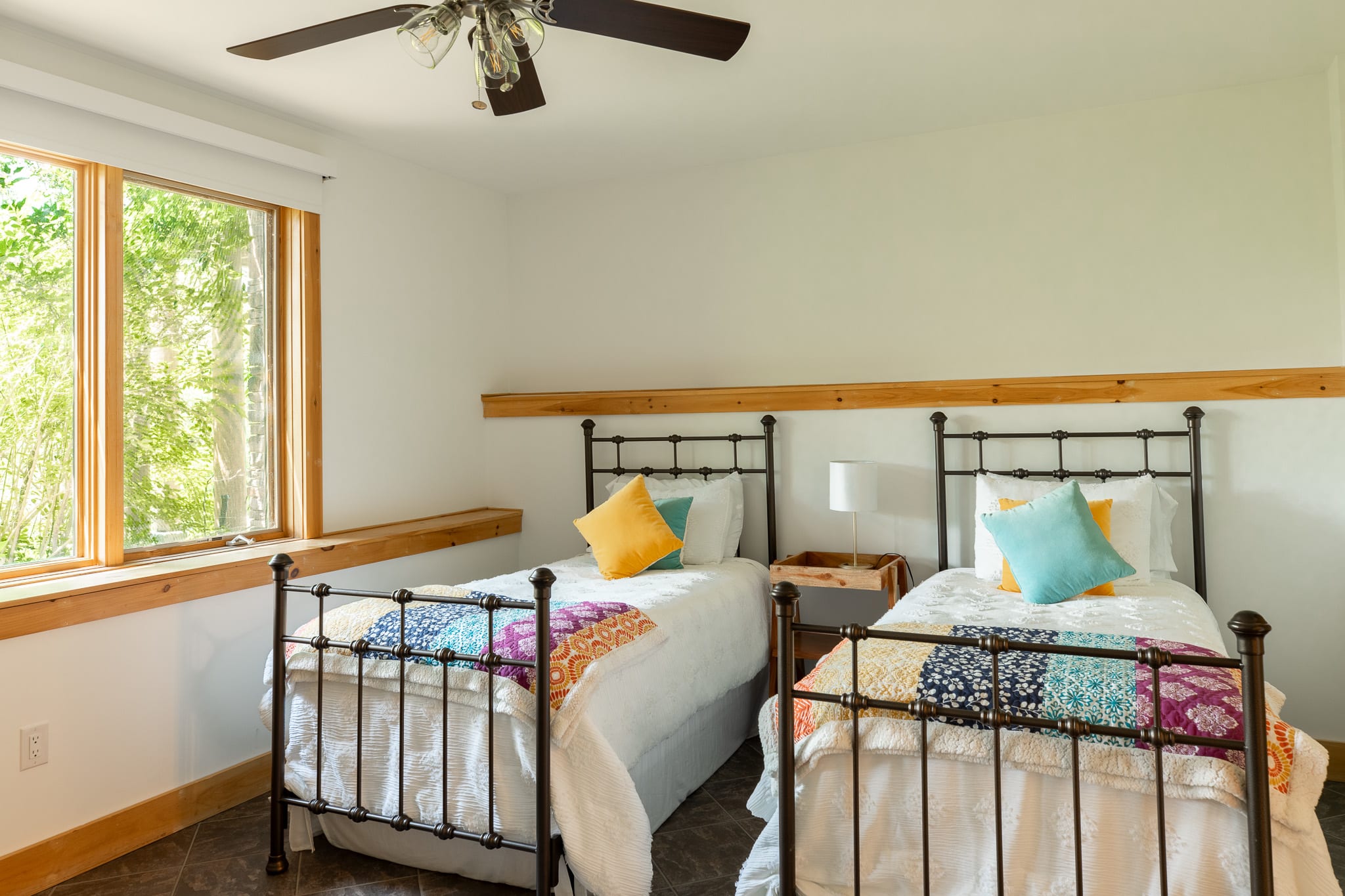 A bright bedroom featuring two twin beds with black metal frames, colorful quilted blankets, and yellow and teal throw pillows, set against white walls with a natural wood trim and large windows overlooking greenery.