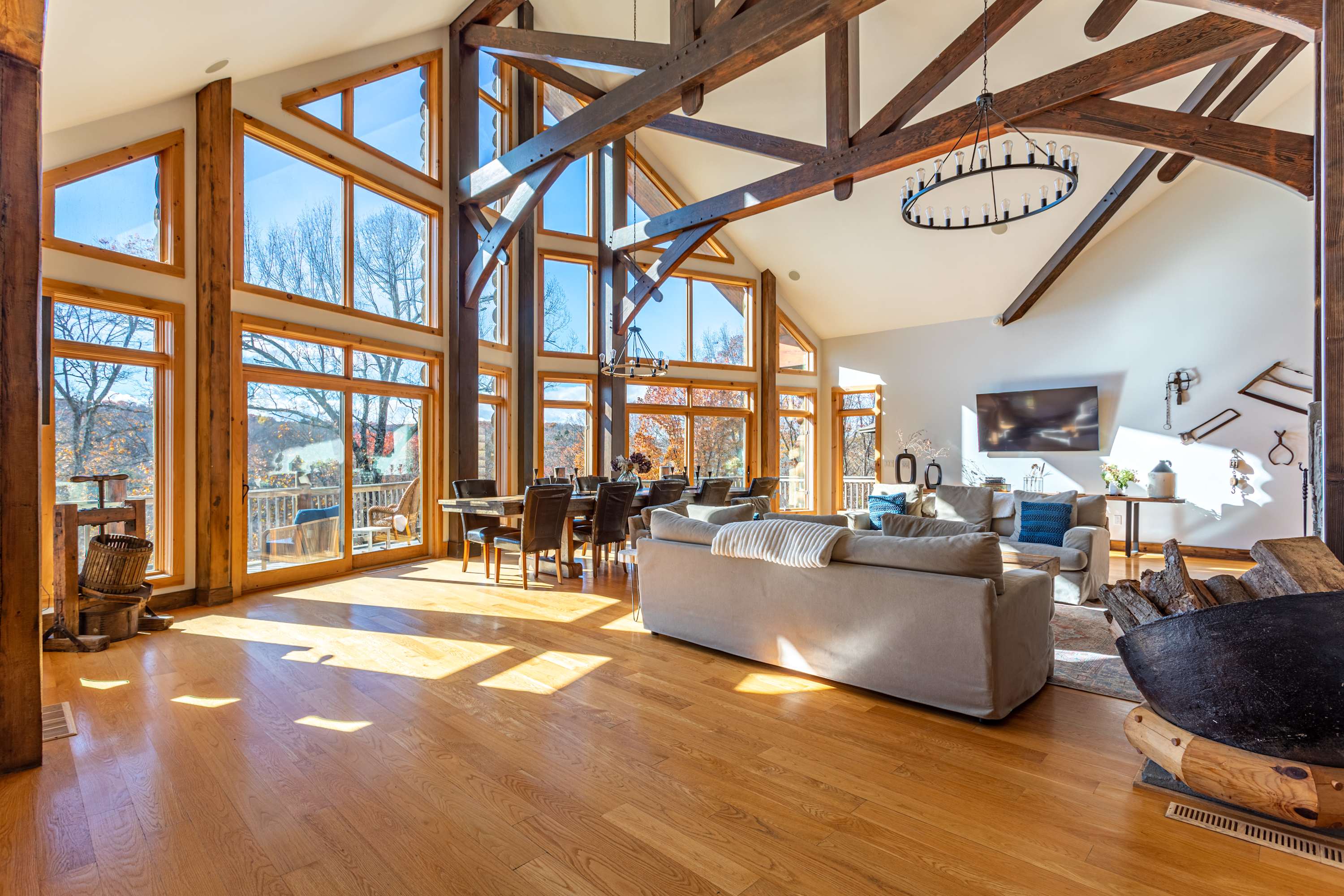 A bright and airy living and dining space with soaring vaulted ceilings, prominent exposed wooden beams, and a wall of large windows showcasing the scenic outdoor landscape.