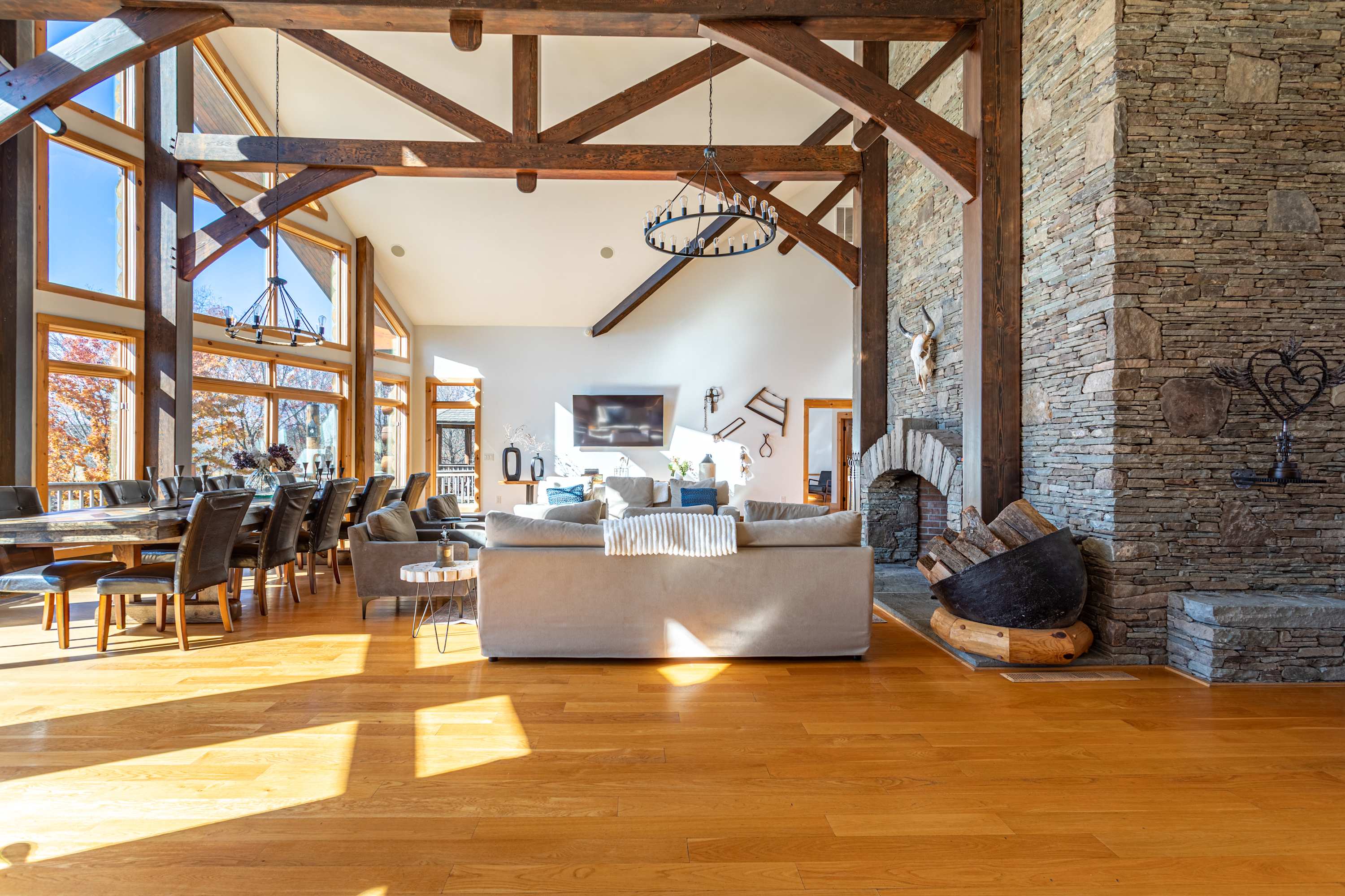 A grand living space with a towering stone fireplace, exposed wooden beams, and an open layout leading to a dining area with large windows overlooking the outdoors.