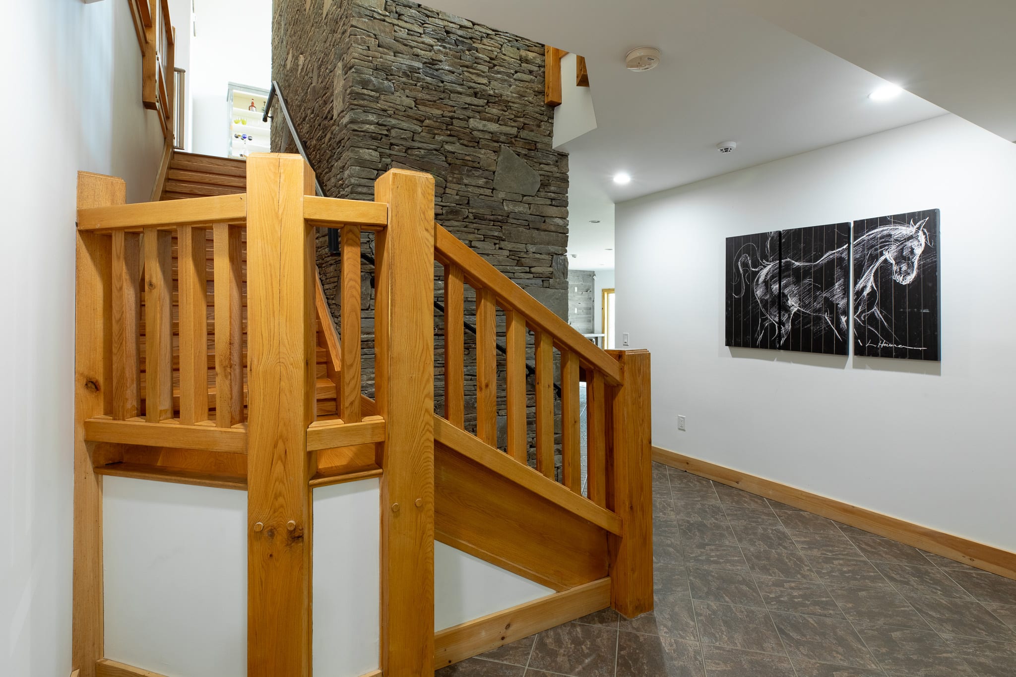 A well-lit foyer featuring a natural wood staircase with a sturdy railing, a stone accent wall, and dark tiled flooring, with a large black and white horse artwork on the wall, leading to other areas of the house.