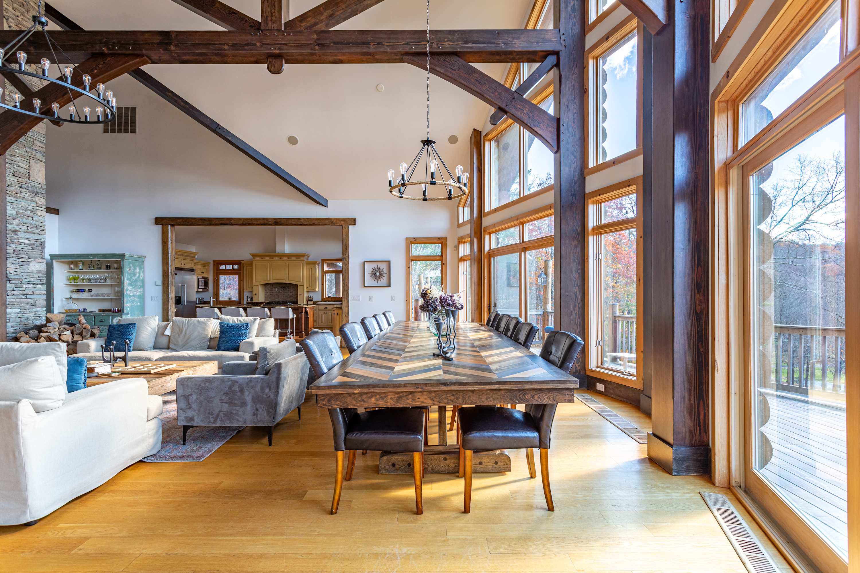 A long, rustic wooden dining table with a herringbone pattern top, surrounded by dark leather chairs, positioned in front of expansive windows that offer bright outdoor views, under a modern chandelier.