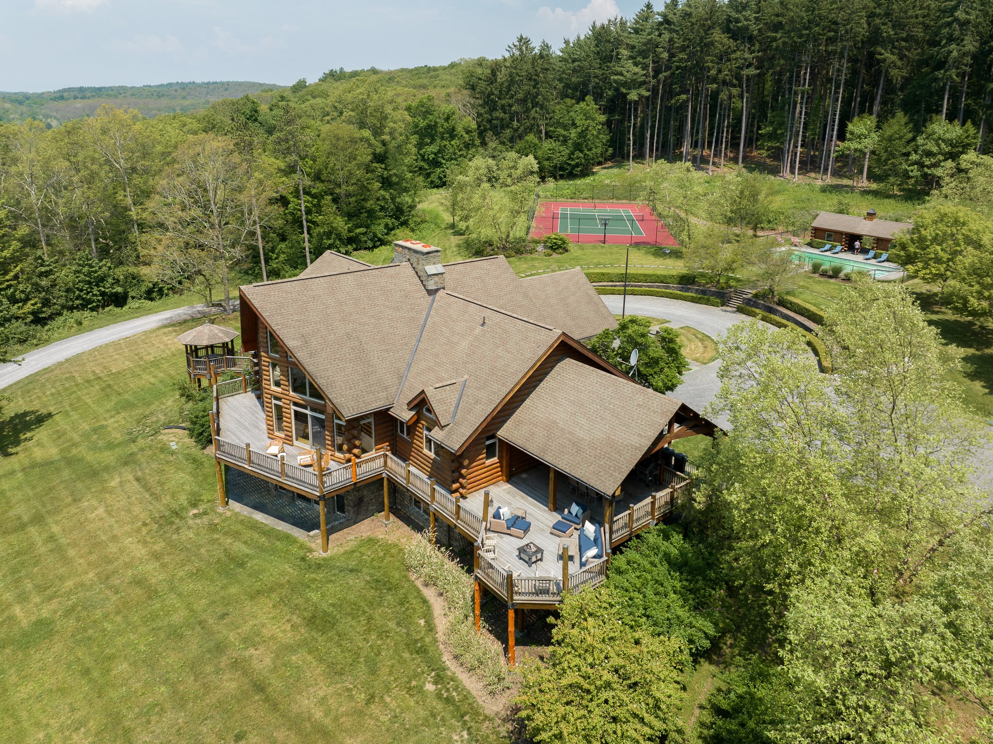 A wide aerial shot of the expansive log cabin property, featuring the main house with its large deck, a circular driveway, a tennis court, and a swimming pool, all nestled within a vast wooded landscape.