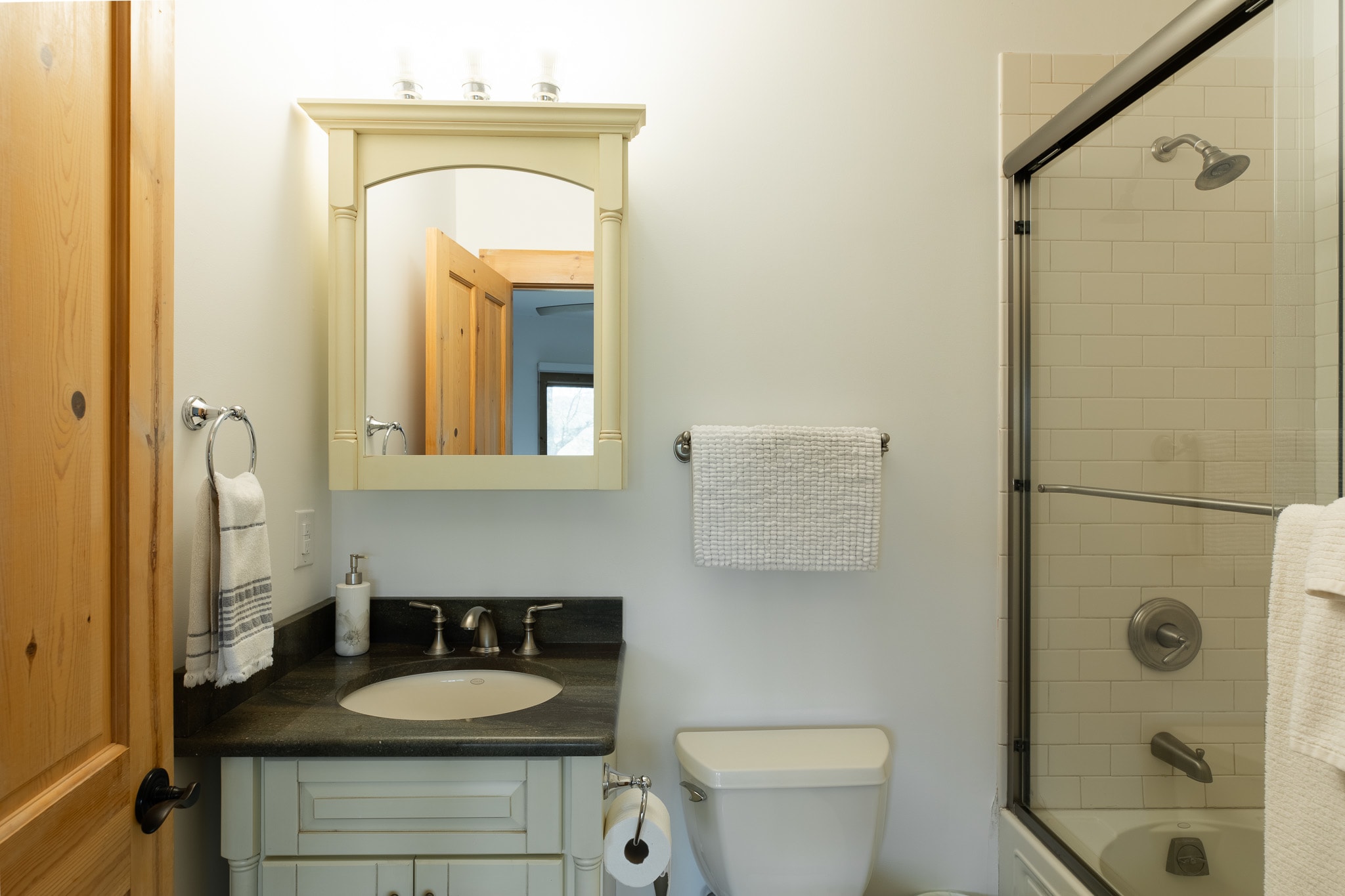 A bright bathroom featuring a light-colored vanity with a dark countertop, a toilet, and a shower/tub combo with subway tile surround and a glass enclosure. A wooden door is visible on the left.