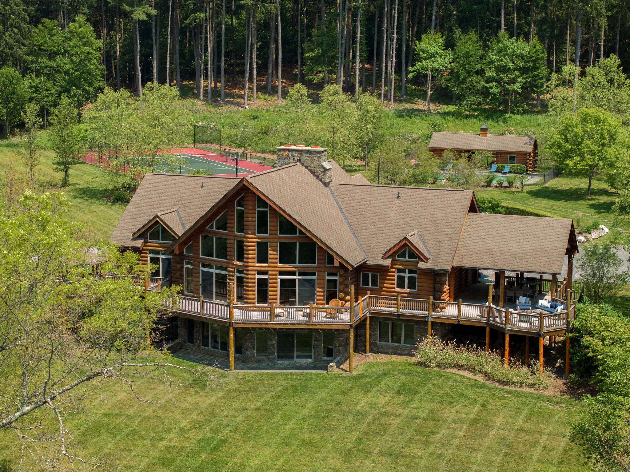 An aerial view of a large log cabin house with a multi-level deck, situated amidst a sprawling green lawn and dense forest, with a tennis court and pool area visible in the distance.