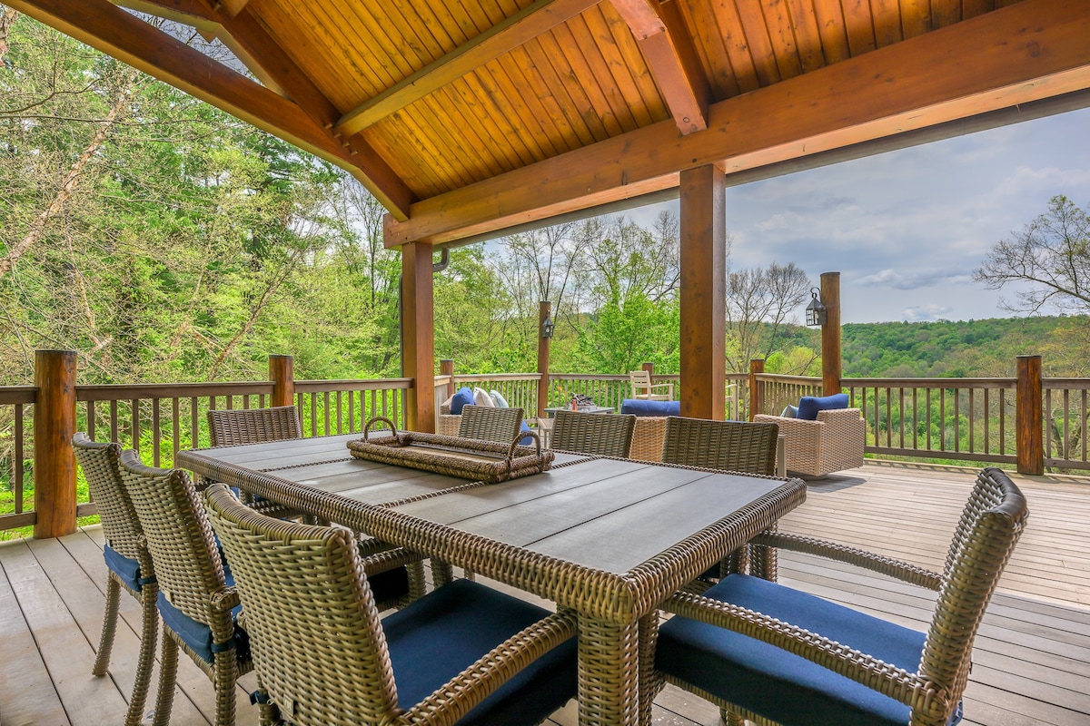 A spacious outdoor deck with a large wicker dining table and chairs, set under a wooden pergola, offering expansive views of a lush, tree-filled valley. Additional outdoor seating is visible in the background.