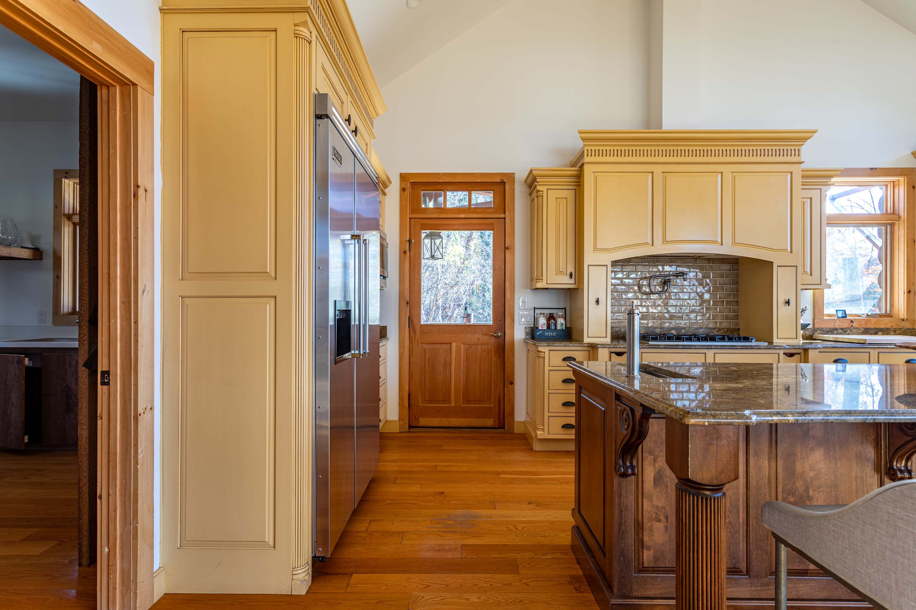 View of the kitchen's stainless steel refrigerator and light yellow pantry cabinets, with a wooden door leading outside and a glimpse of the gas range area.