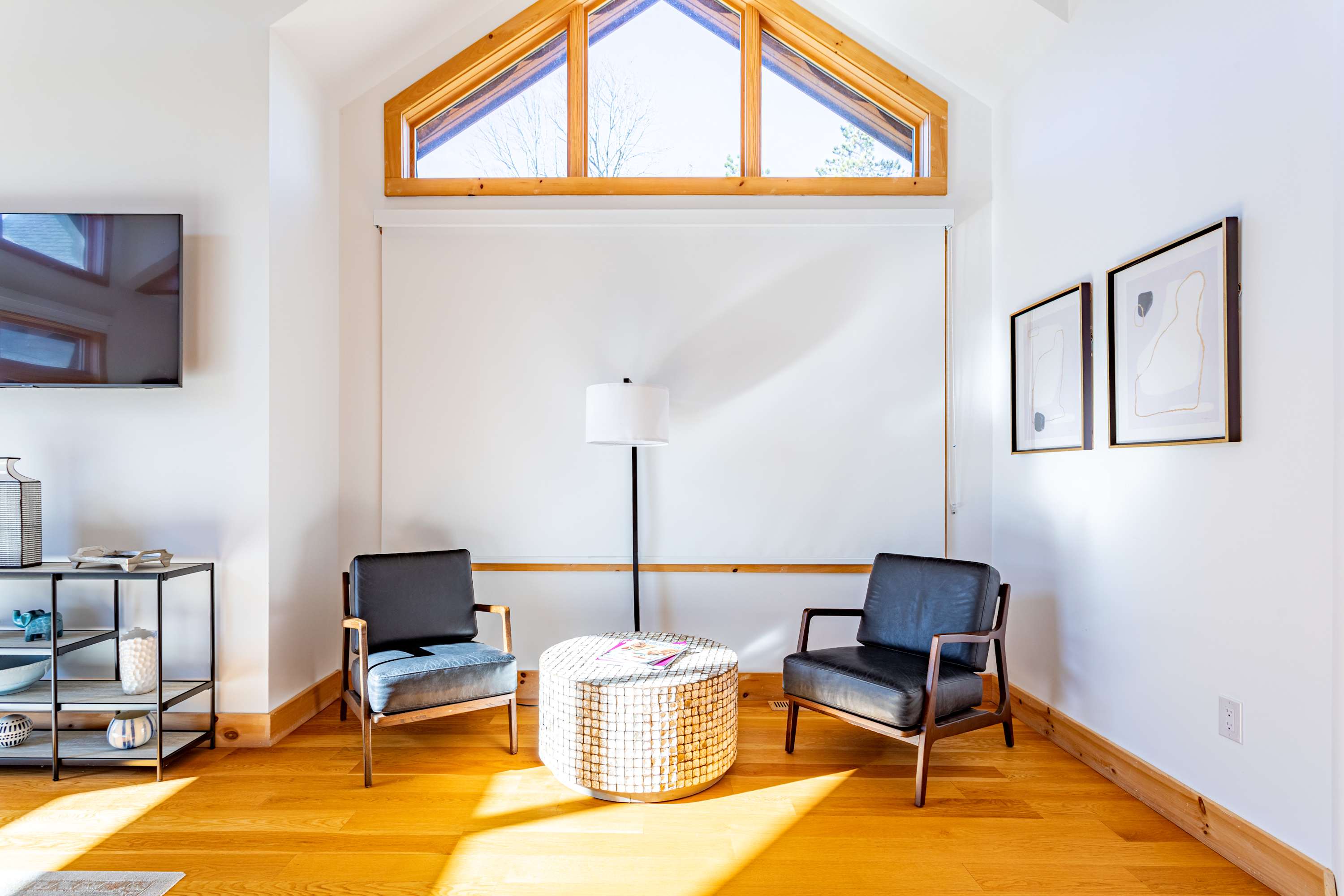 A bright, cozy seating nook with two dark armchairs and a round woven coffee table, positioned in front of a large window with an arched top, allowing ample natural light into the room.