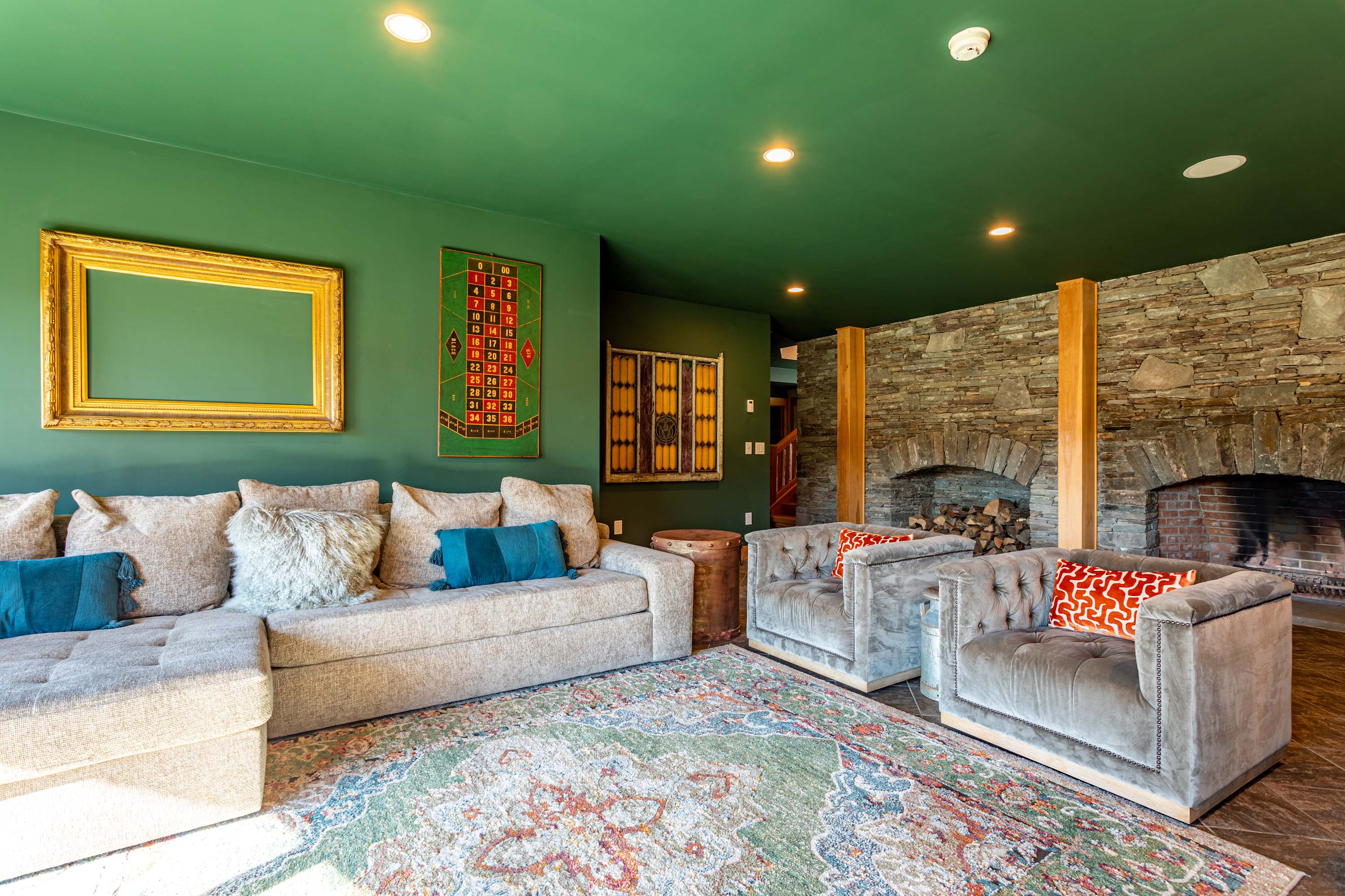 A cozy living area with a large sectional sofa and two armchairs, centered around a colorful patterned rug. The room features a striking green ceiling, a stone fireplace, and a large framed roulette board on the wall.