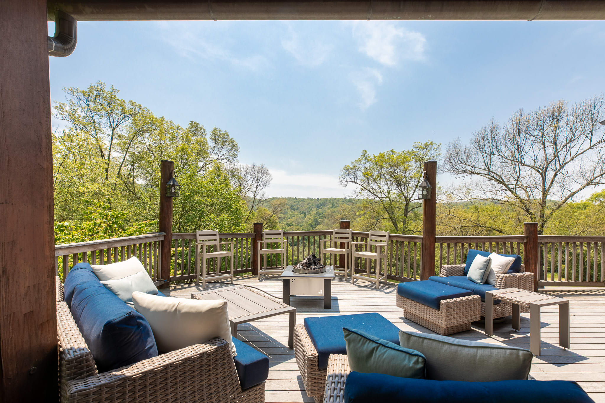 A spacious outdoor deck with multiple seating areas, including wicker sofas with blue cushions and a fire pit, overlooking a lush green valley under a bright sky. The deck features wooden railings and support beams.