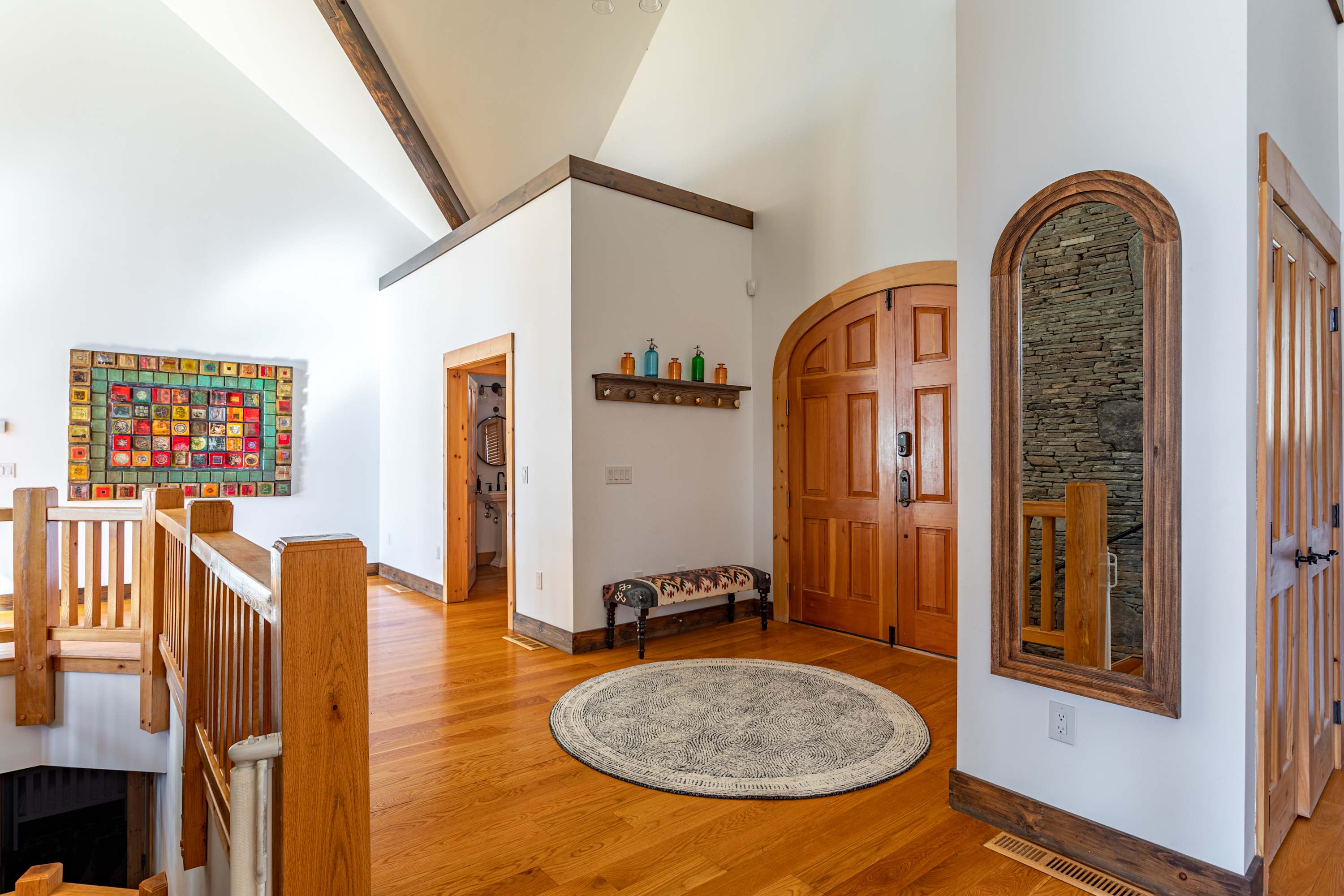 Expansive foyer with light hardwood floors and high vaulted ceilings, featuring a wooden staircase railing leading downstairs, and a colorful square art piece on the wall.