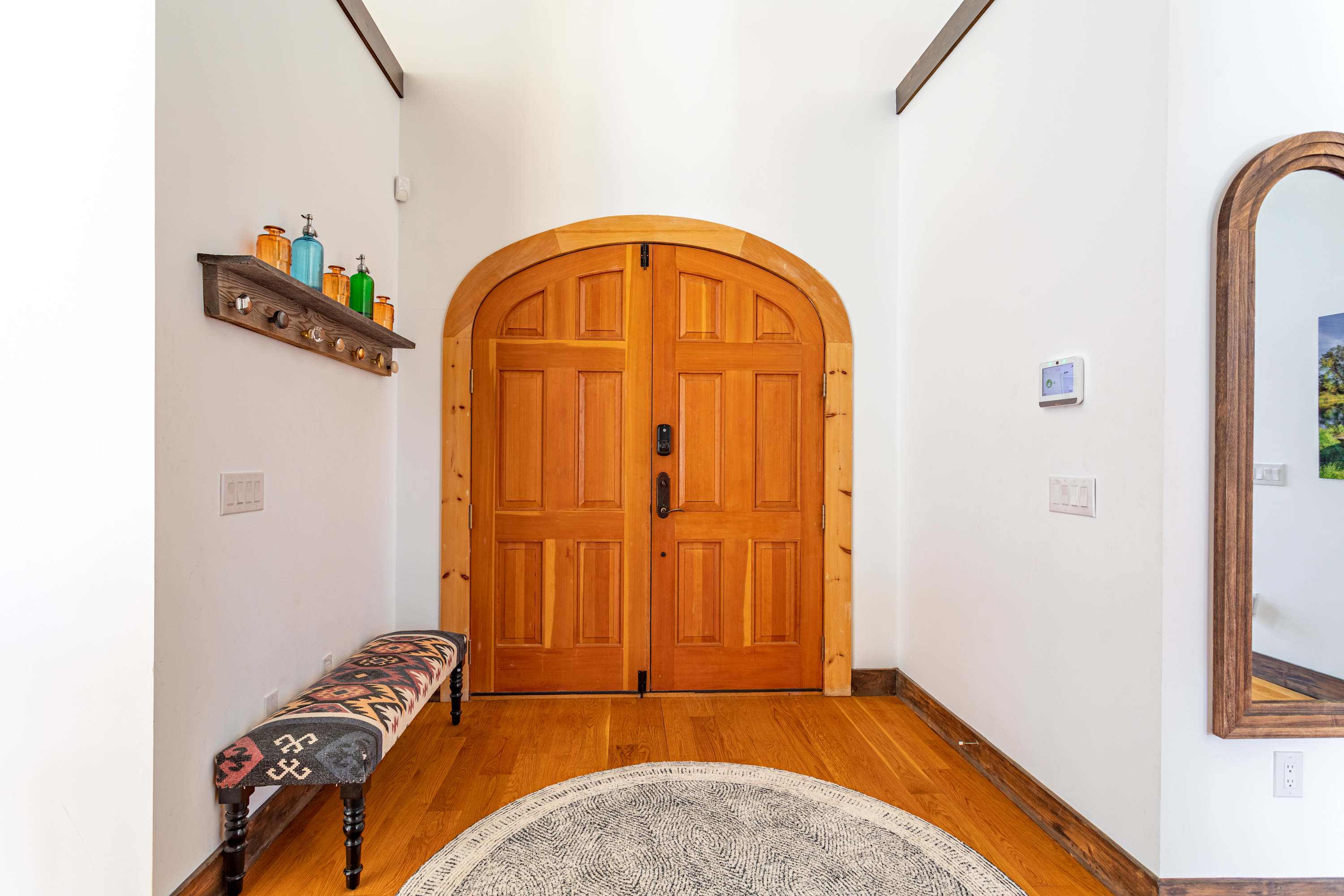 Bright interior foyer with light hardwood floors and white walls, featuring double arched wooden entry doors, a patterned bench, and a decorative shelf with colorful bottles.
