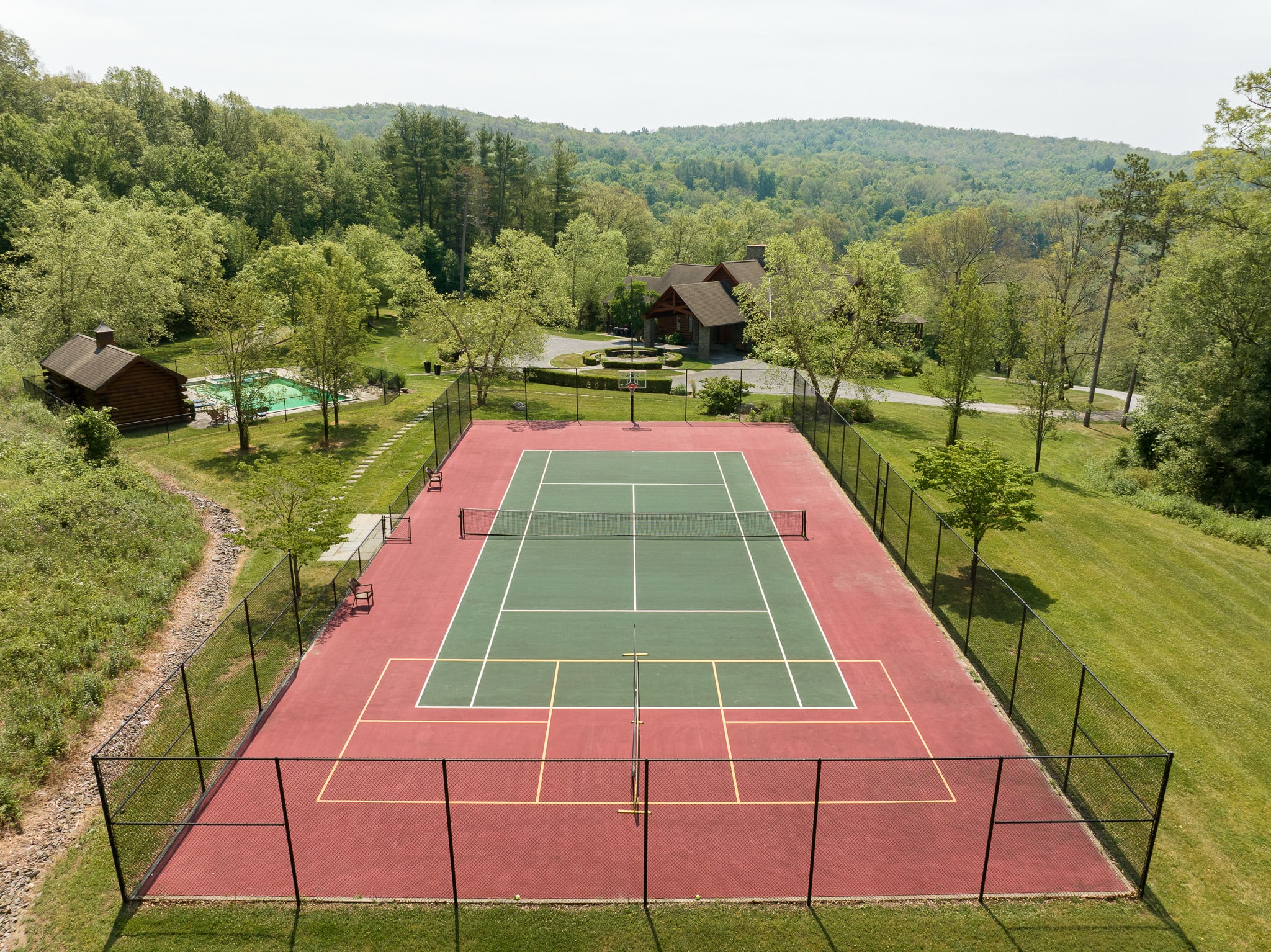 Large outdoor tennis and pickleball court with a red and green surface, enclosed by a black fence, surrounded by green grass and distant trees under a bright sky.