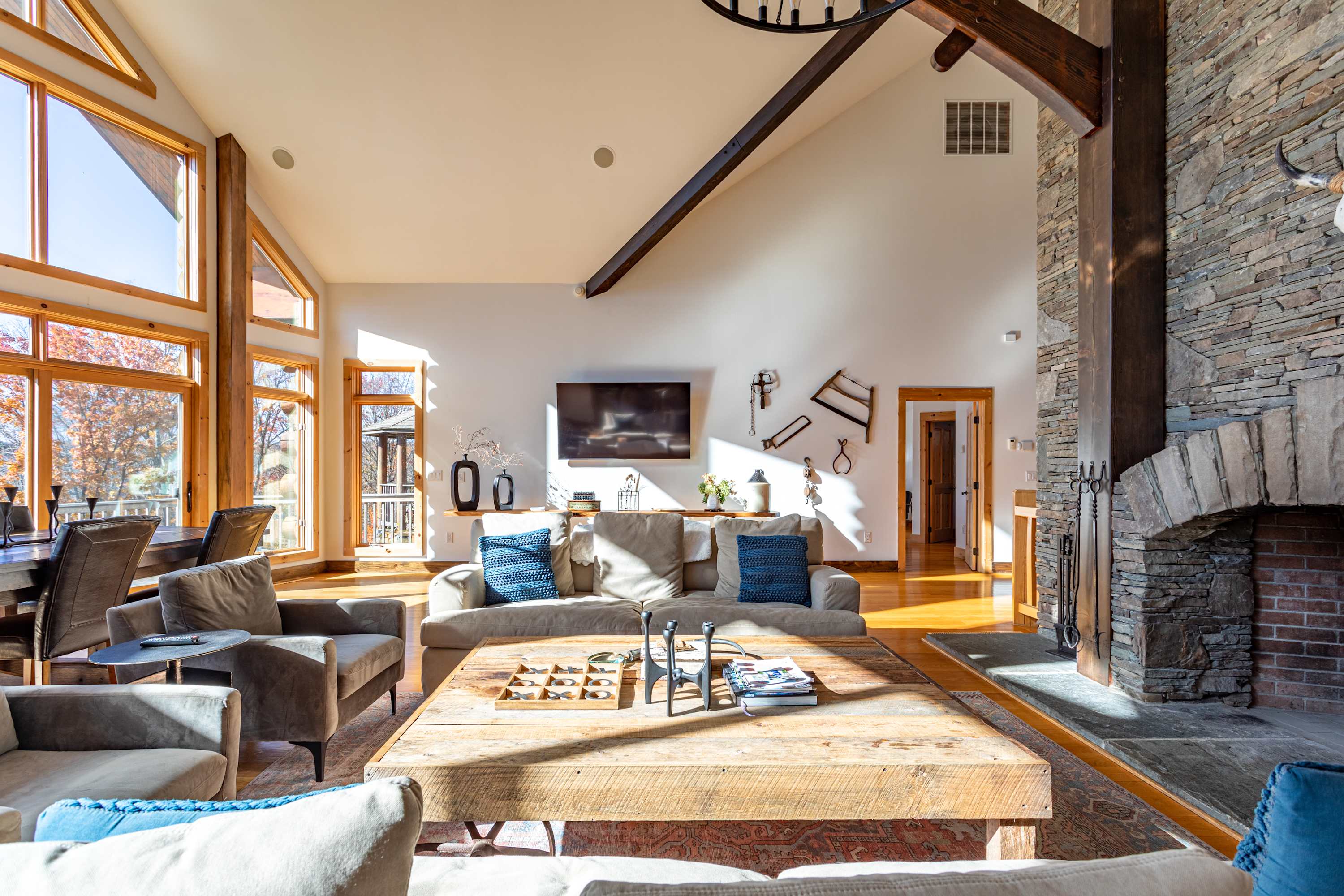 An inviting living room with a large wall-mounted TV, decorative wall art, and comfortable seating, complemented by a grand stone fireplace and expansive windows, all under a vaulted ceiling.