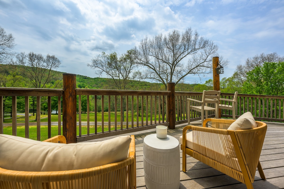 A section of a wooden deck furnished with two woven rattan armchairs with cream cushions and a white cylindrical side table, offering expansive views of a wooded landscape under a partly cloudy sky.