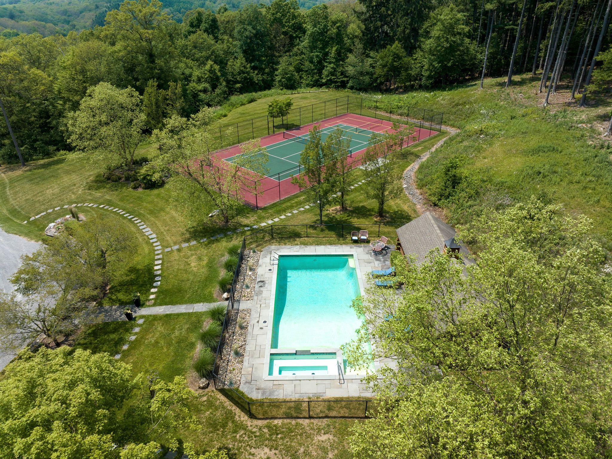 An aerial view of the property showcasing a rectangular swimming pool with a hot tub, a red and green tennis court, and winding pathways, all surrounded by lush green lawns and dense trees.
