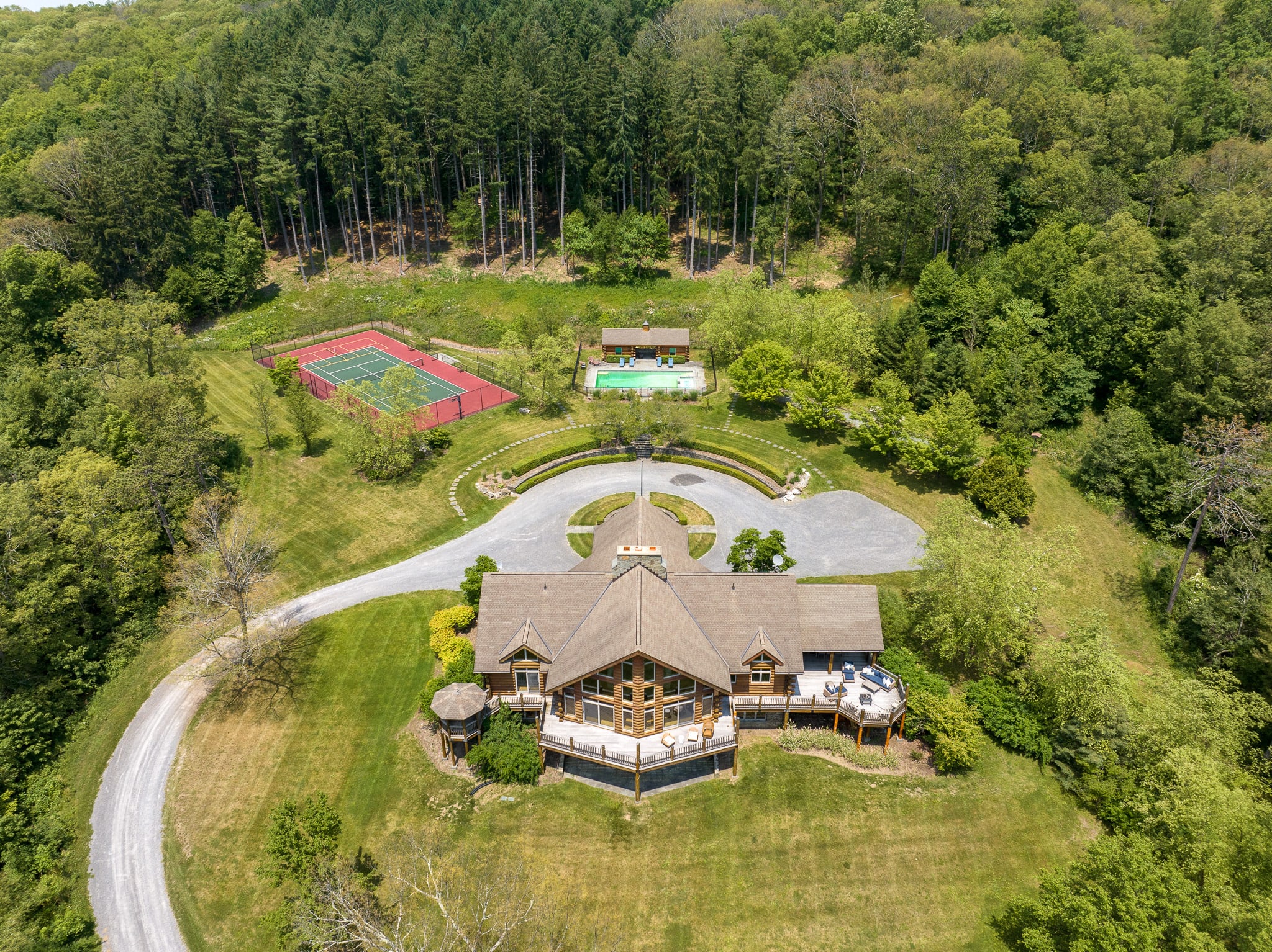 Aerial view of a large log cabin-style house with a circular driveway, surrounded by lush green trees, featuring a tennis court and a pool house with a swimming pool.