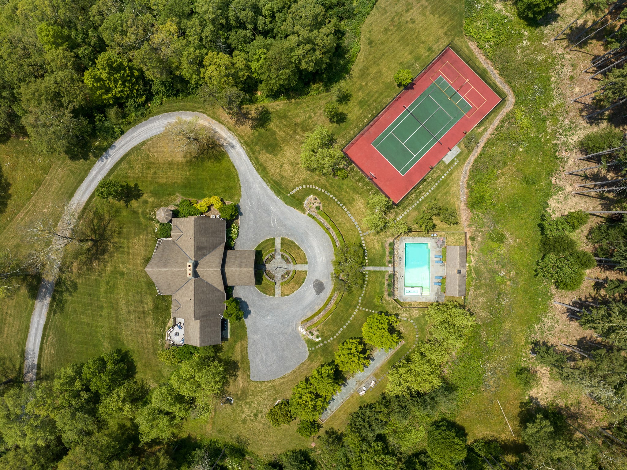 An expansive aerial view showcases a large property featuring a multi-winged house, a circular gravel driveway, a bright blue swimming pool, and a red tennis court, all nestled within dense green woodlands.