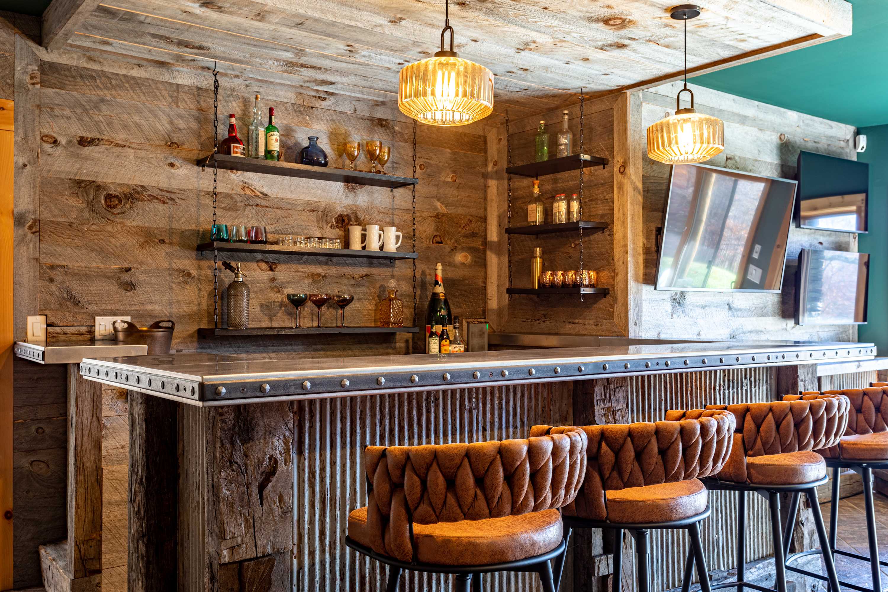 A detailed view of the rustic bar's metal countertop and the reclaimed wood back wall, adorned with open shelves holding various bottles and glassware. Two amber-toned pendant lights illuminate the area, with a large TV visible to the right.