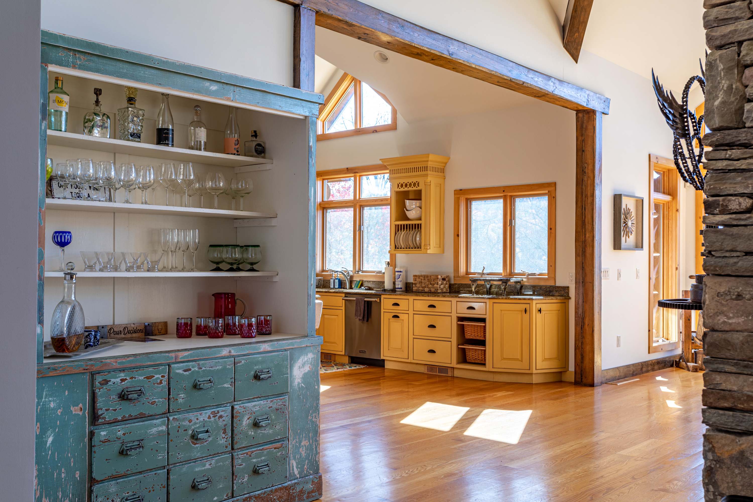 A rustic kitchen featuring light yellow cabinetry, granite countertops, and a large distressed teal hutch filled with glassware, all set against light hardwood floors and vaulted ceilings with exposed wooden beams.