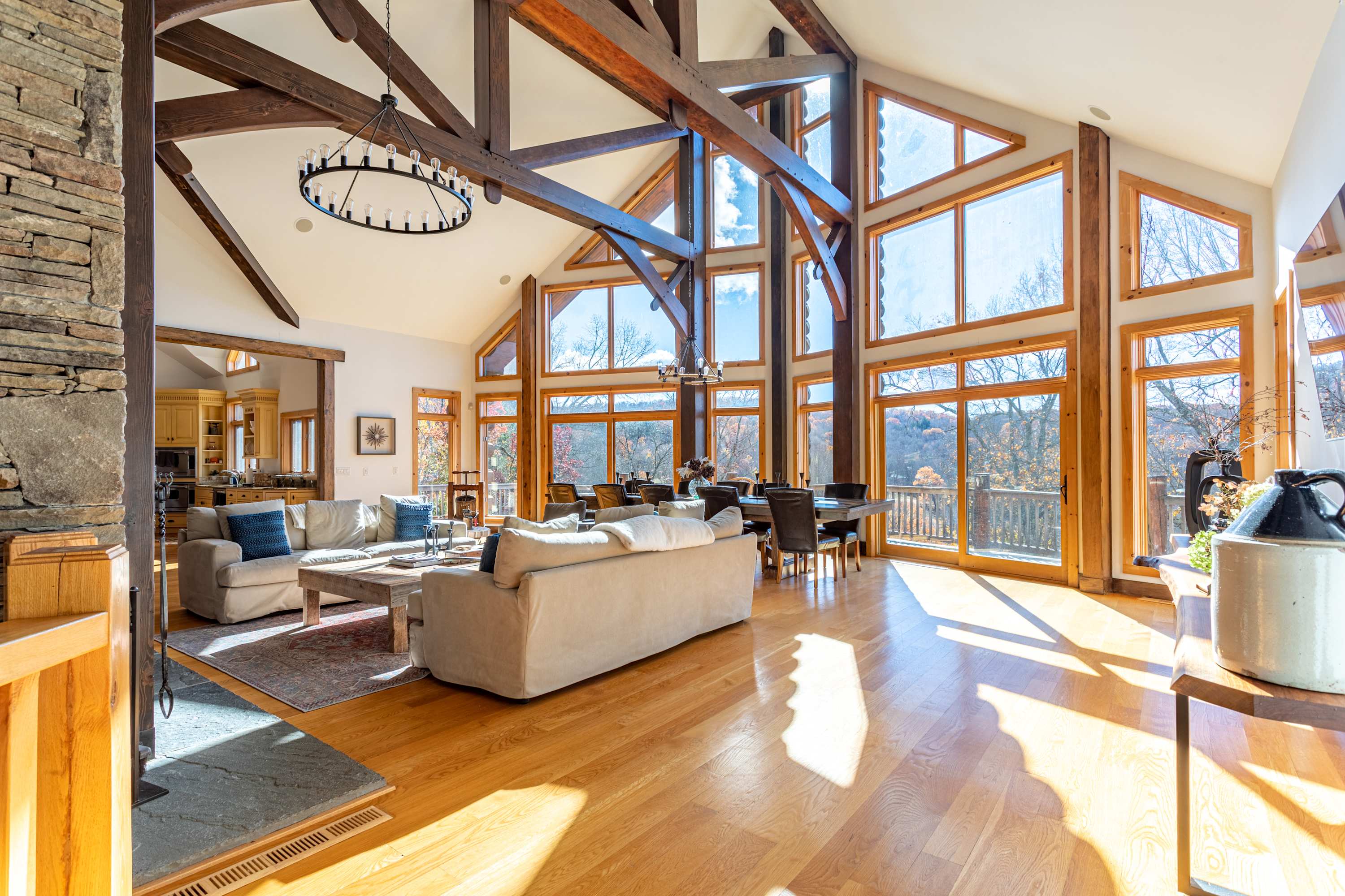 A spacious open-concept living and dining area with light hardwood floors, high vaulted ceilings, and large windows, featuring a comfortable seating arrangement and a long dining table, with the kitchen visible in the background.