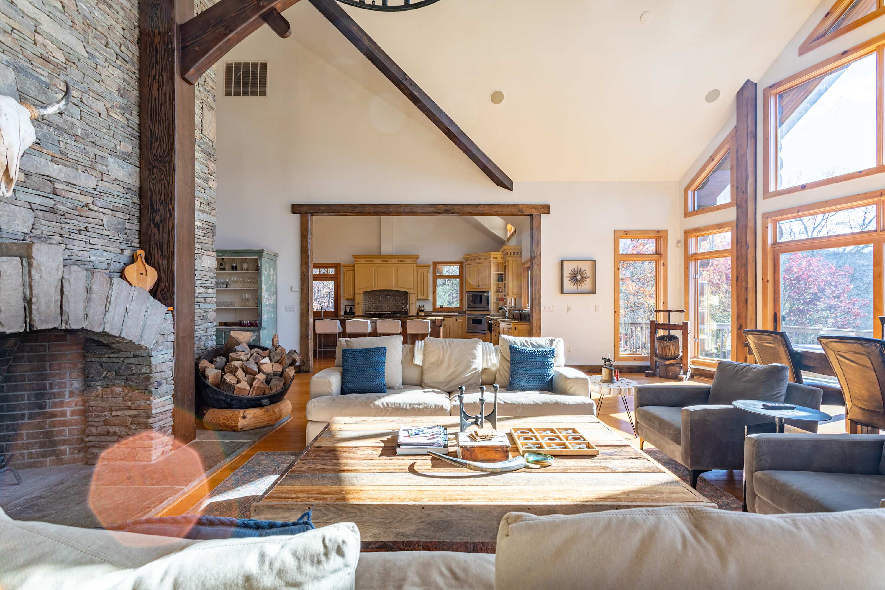 A wide shot of the spacious living room, showcasing the stone fireplace, light-colored sofas, and a large wooden coffee table, with a glimpse of the kitchen and large windows offering natural light.