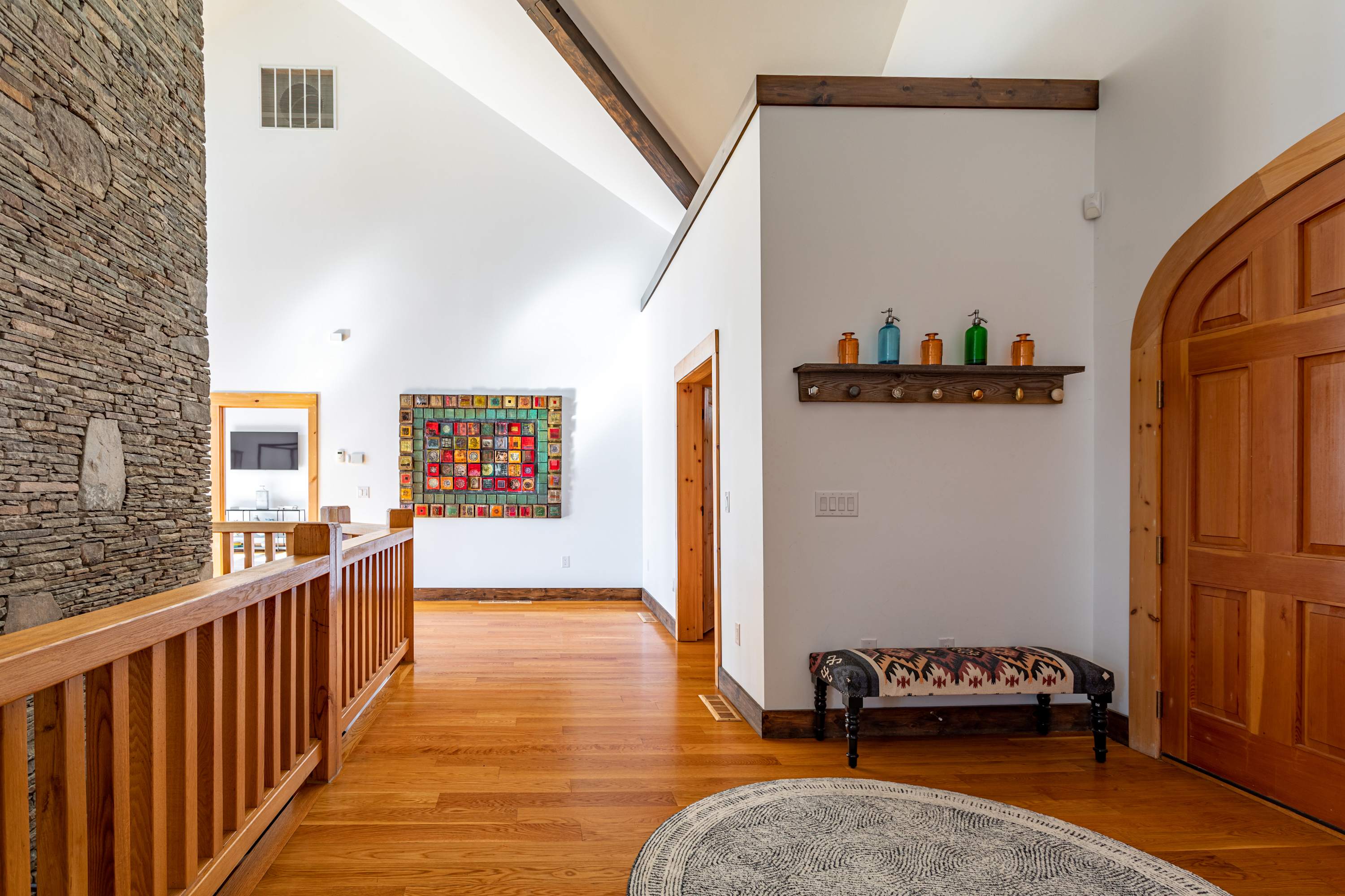 Bright hallway with hardwood floors and white walls, showcasing a large colorful square art piece and a rustic stone wall on the left, with a wooden railing indicating a staircase.