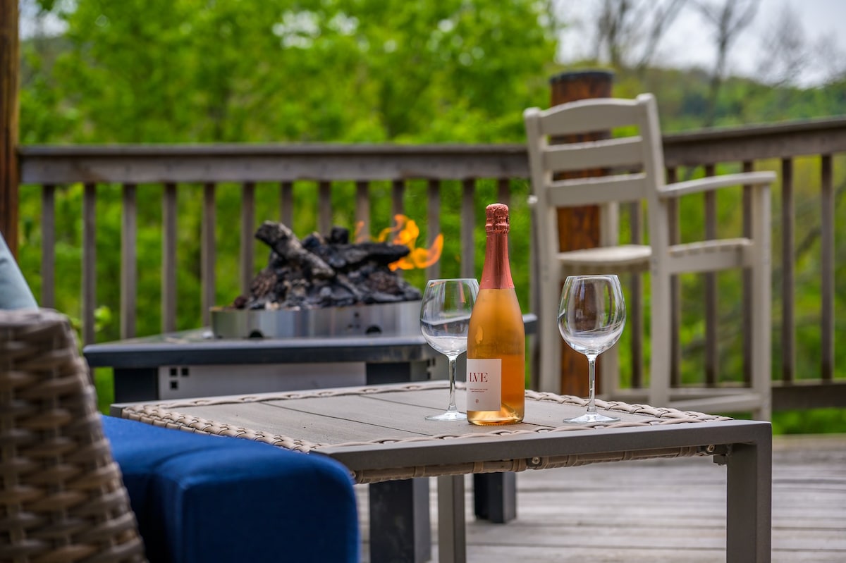 A close-up view of a modern fire pit with burning logs on a wooden deck, flanked by a bottle of rosé wine and two wine glasses on a dark outdoor coffee table. Green trees are visible in the background.