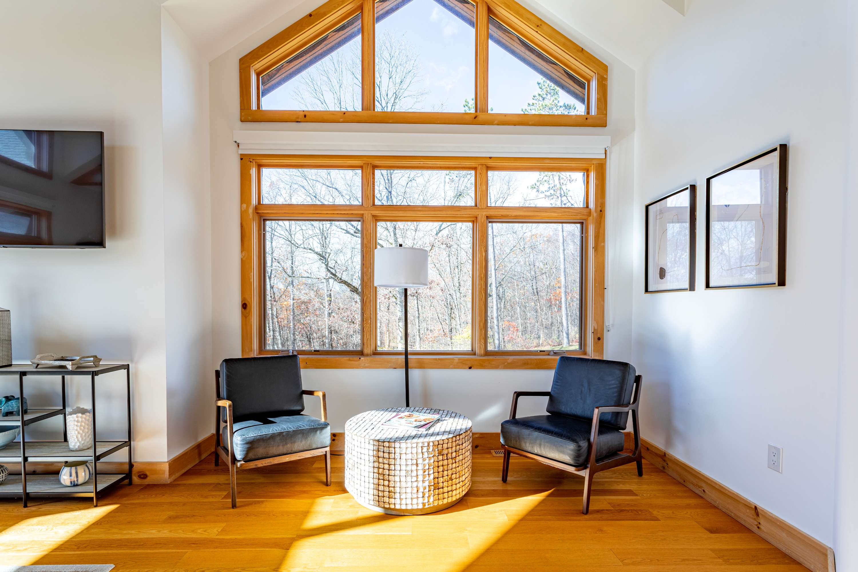A bright seating area featuring two dark armchairs and a round woven coffee table, set against a backdrop of large windows with an arched top, offering views of the surrounding trees.