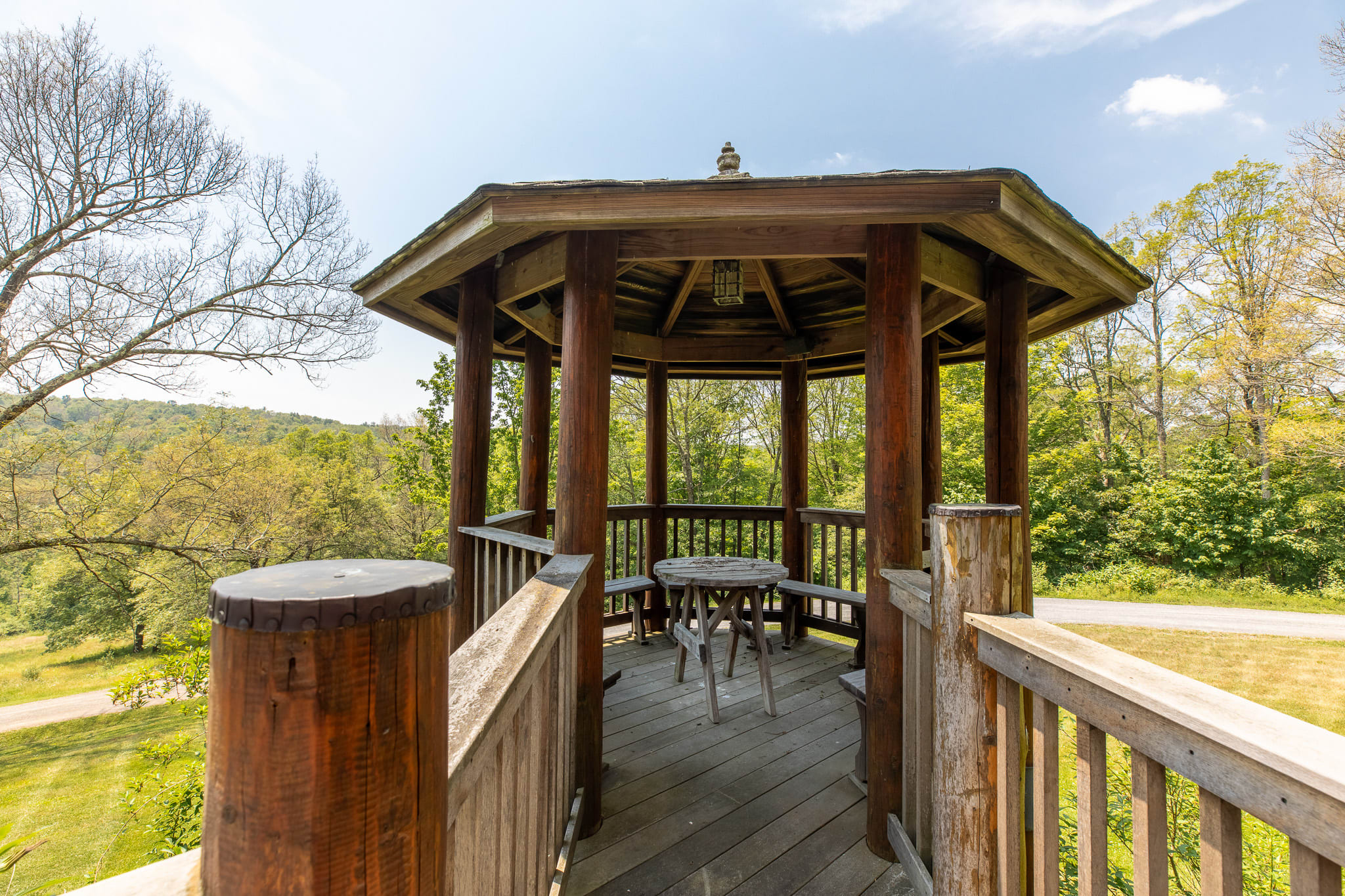 A rustic wooden gazebo with a hexagonal roof and open sides, featuring a small round wooden table and benches, situated on a raised deck overlooking a winding path and green landscape.
