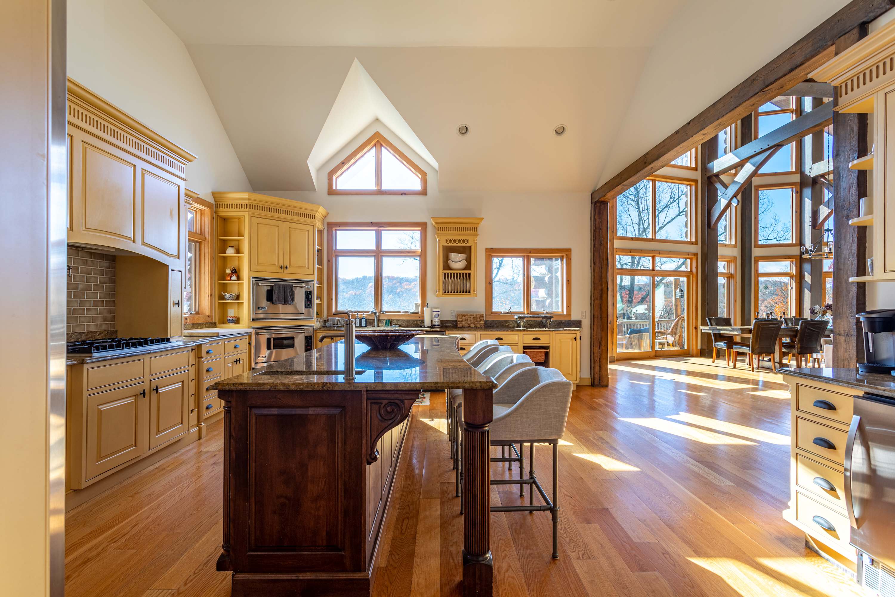 Expansive kitchen with a large wooden island, light yellow cabinets, stainless steel appliances, and a dining area visible through a wide opening with large windows and exposed beams.
