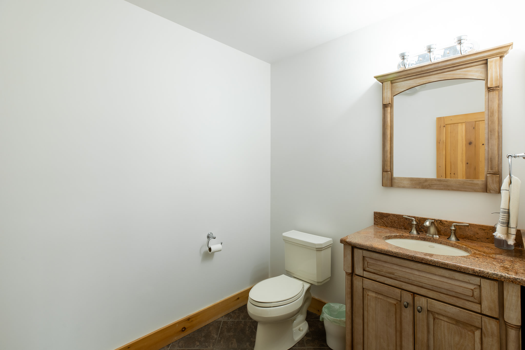 A clean bathroom with a white toilet, a wooden vanity with a granite countertop and sink, and a framed mirror above, all set against white walls with a natural wood baseboard.