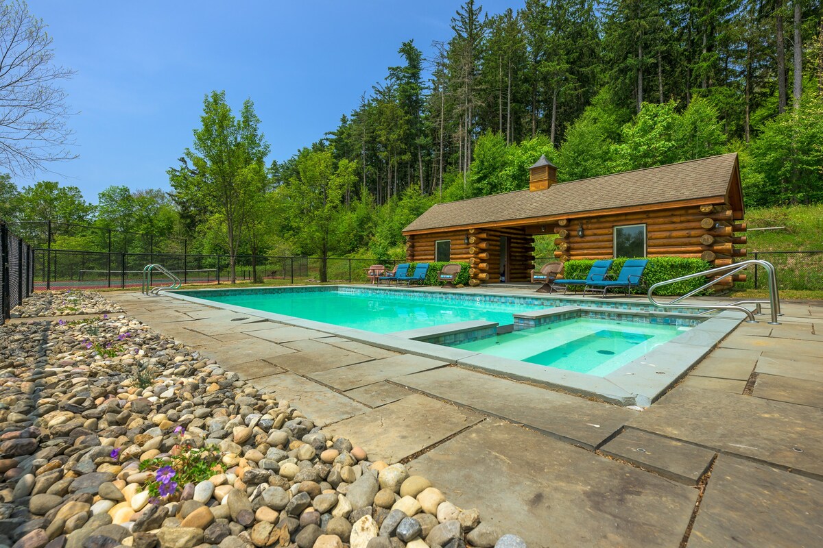 A sparkling blue rectangular swimming pool with an integrated hot tub, surrounded by stone paving and decorative rocks, with a rustic log cabin pool house and lush green trees in the background.
