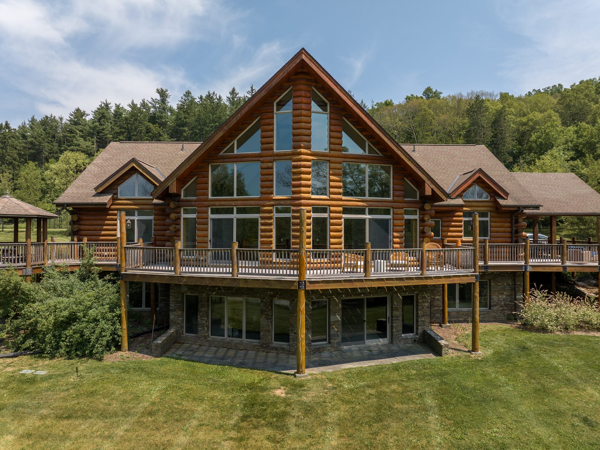Rear view of the large log cabin house with multiple levels, extensive wooden decks, and numerous windows, set against a backdrop of green trees and a clear blue sky.