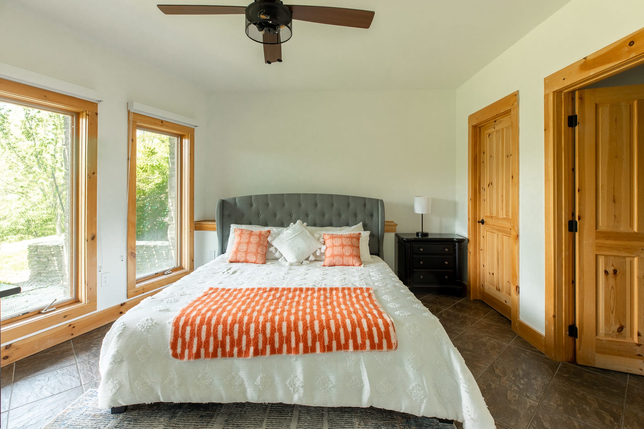 A different angle of the bedroom showing the grey upholstered bed with white and orange bedding, large windows with natural wood trim, and two wooden doors, suggesting an en-suite or closet access.