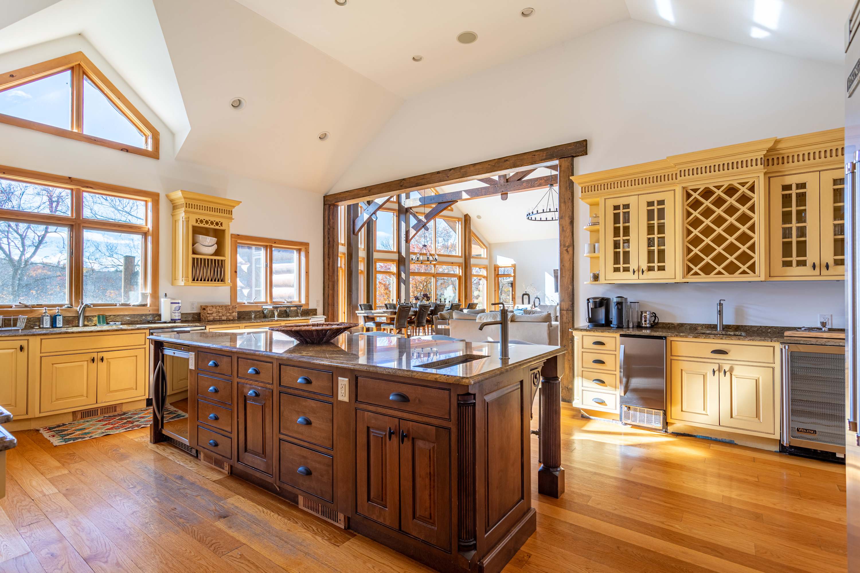 A spacious open-concept kitchen with light yellow perimeter cabinets, a large dark wood island with a granite countertop, and warm hardwood floors. The room features vaulted ceilings and large windows, with a living area visible through a wooden beam archway.