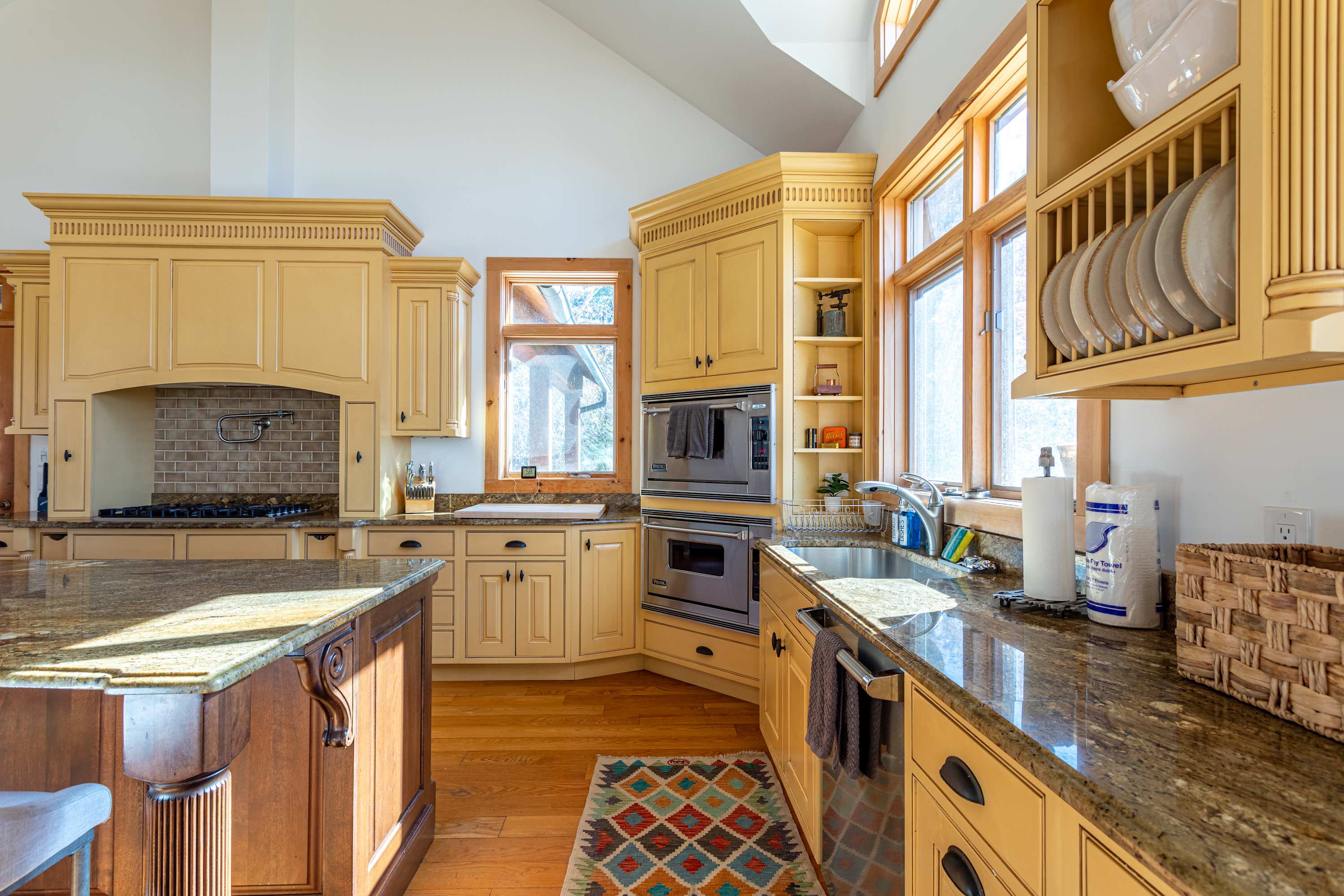 Corner view of the kitchen showcasing light yellow cabinetry, granite countertops, a double oven, and a sink under a window, with a colorful patterned rug on the hardwood floor.
