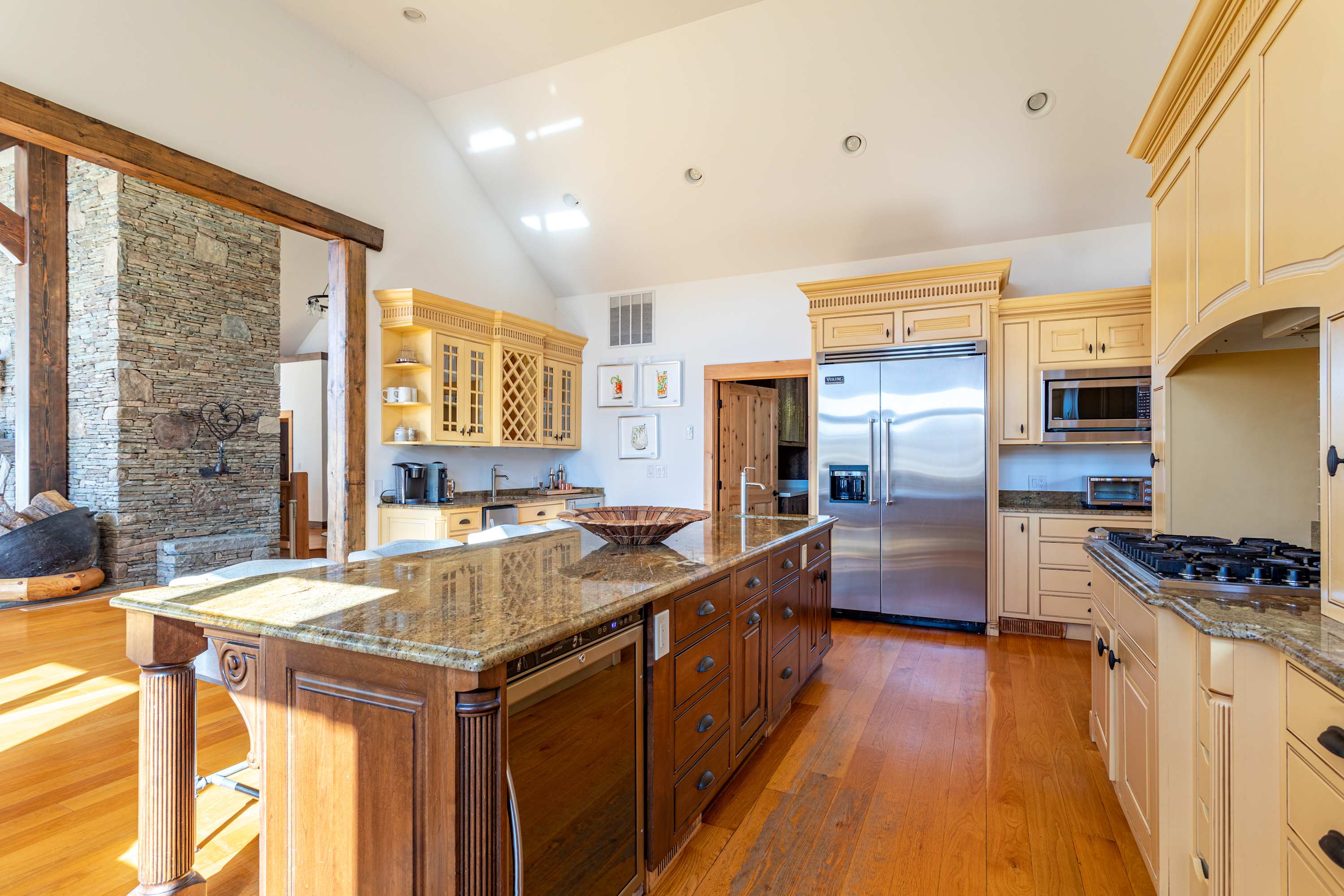 Wide view of a kitchen with light yellow cabinets, a large wooden island with granite countertop, stainless steel refrigerator, and a stone accent wall visible in the adjacent living area.