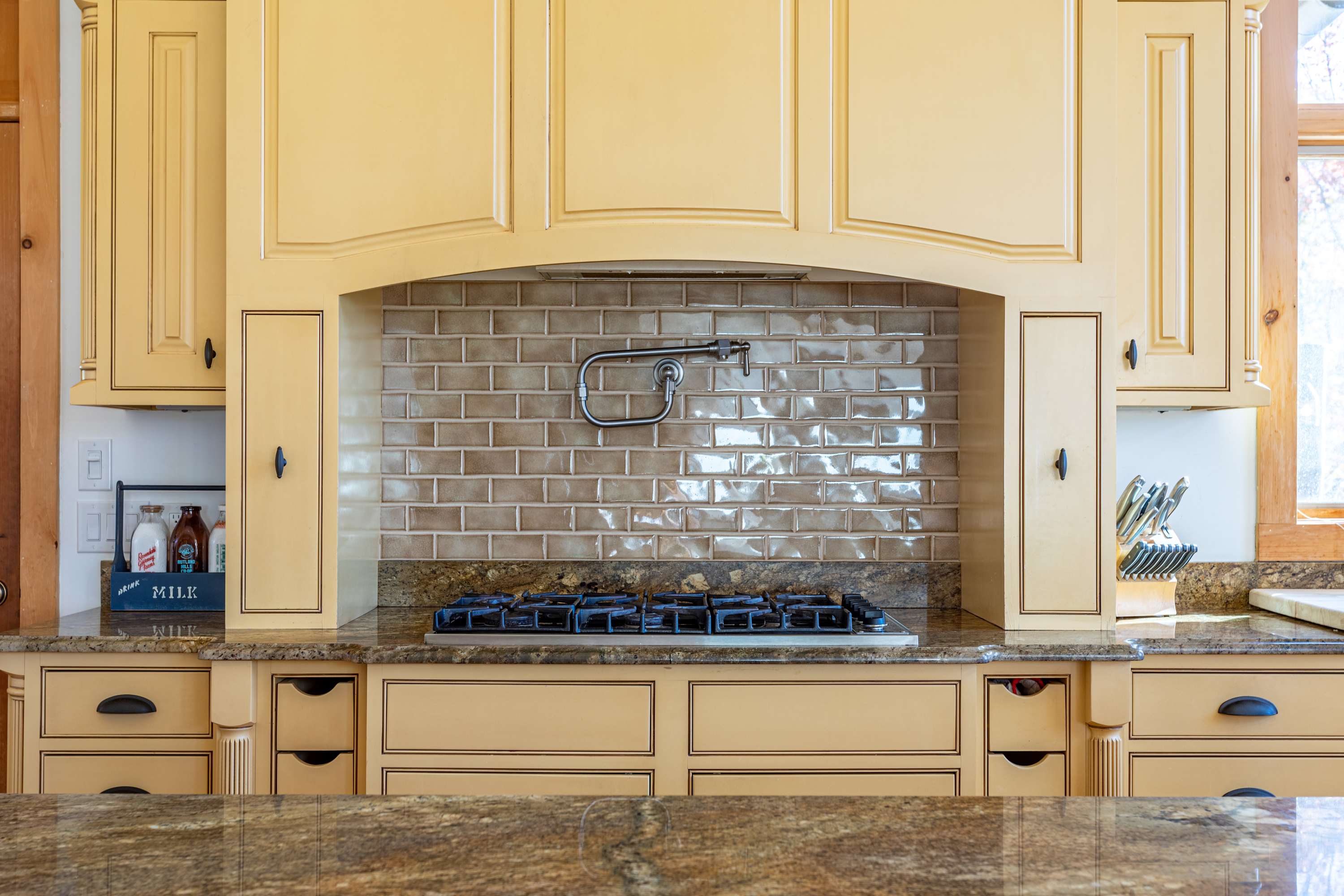 Detailed view of the kitchen's gas range with a pot filler, featuring a beige subway tile backsplash and light yellow cabinetry with black hardware.