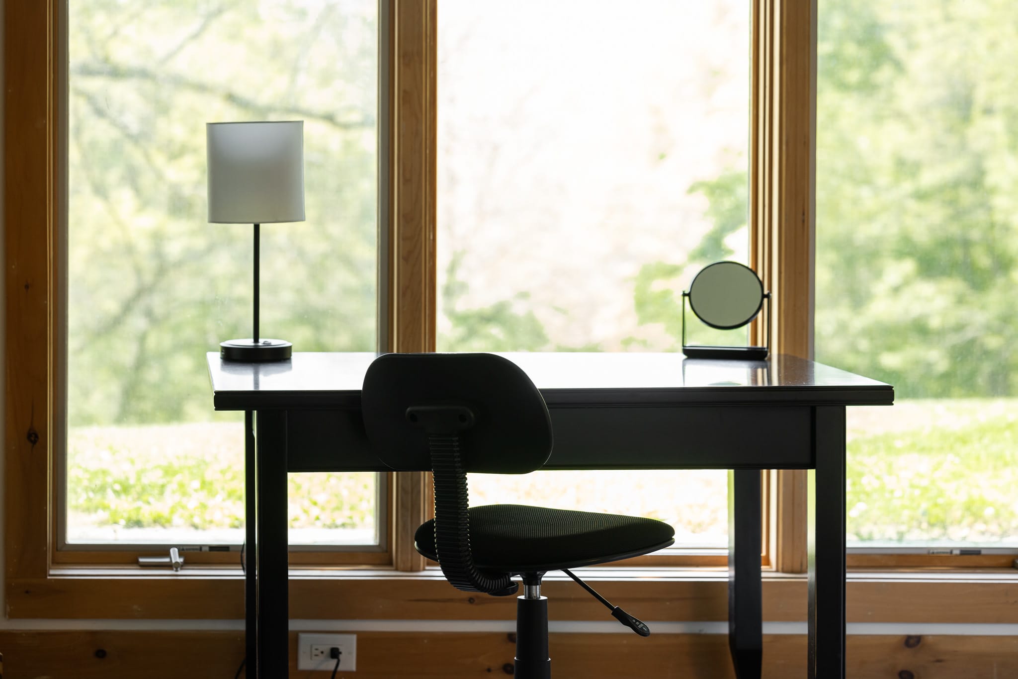 A simple black desk with a task chair, a white lamp, and a small round mirror, positioned in front of large windows offering a bright view of lush greenery. The room features light wood paneling on the lower wall.