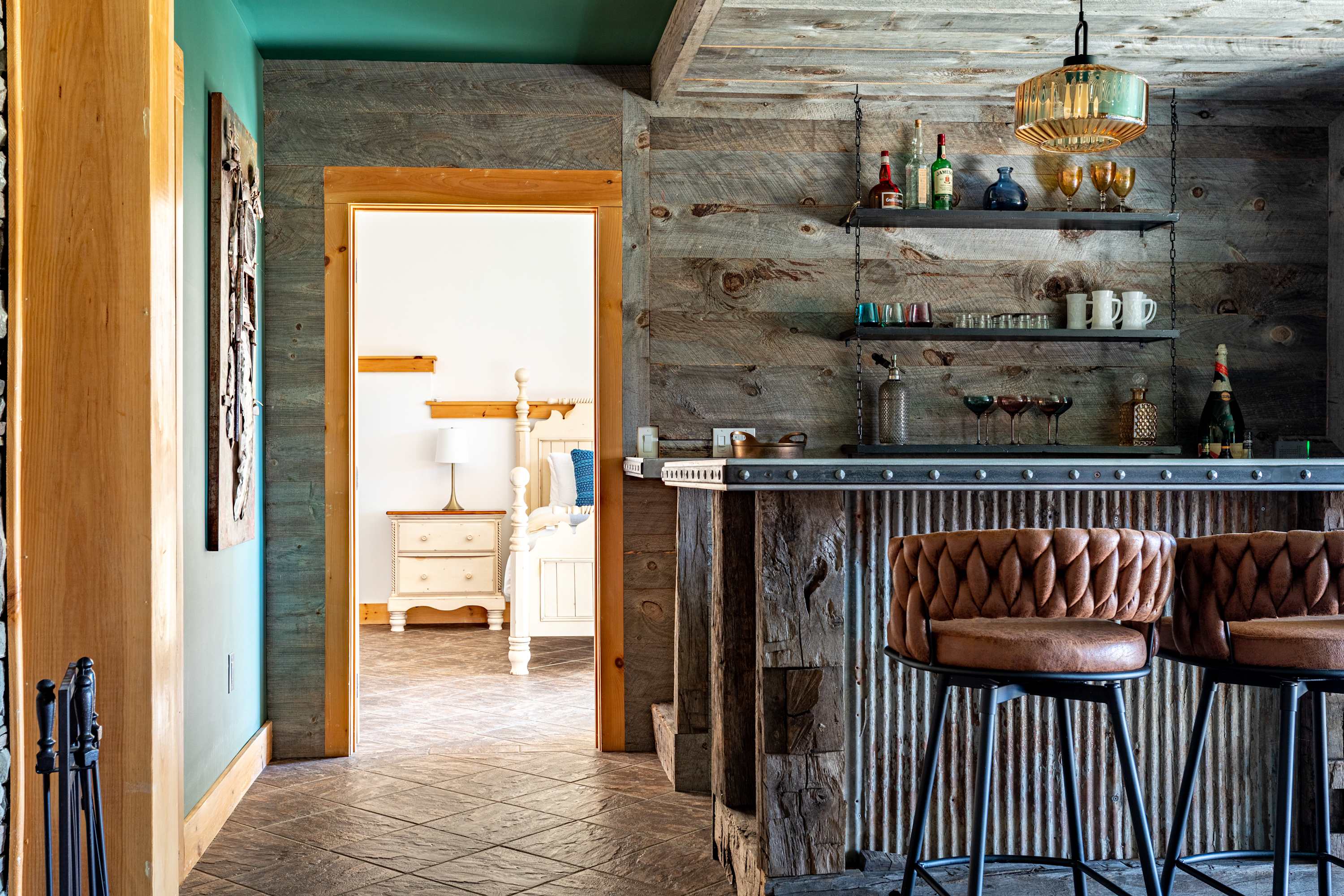 A close-up of the rustic bar area, showcasing the reclaimed wood and corrugated metal construction, with two brown leather bar stools. A doorway to the left reveals a glimpse of a bedroom with a white bed frame and nightstand.